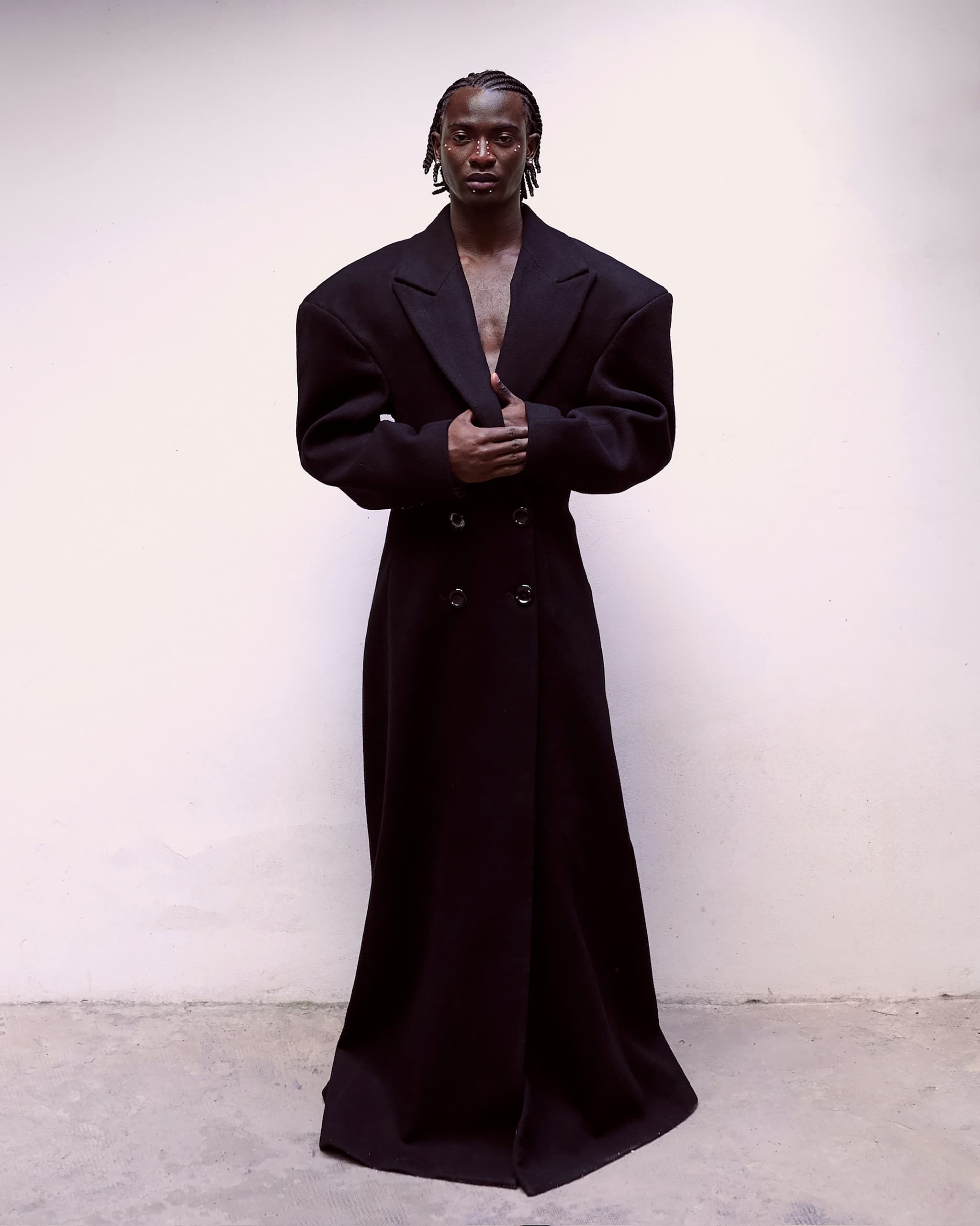 Person wearing an oversized black coat, standing against a minimalist white background.