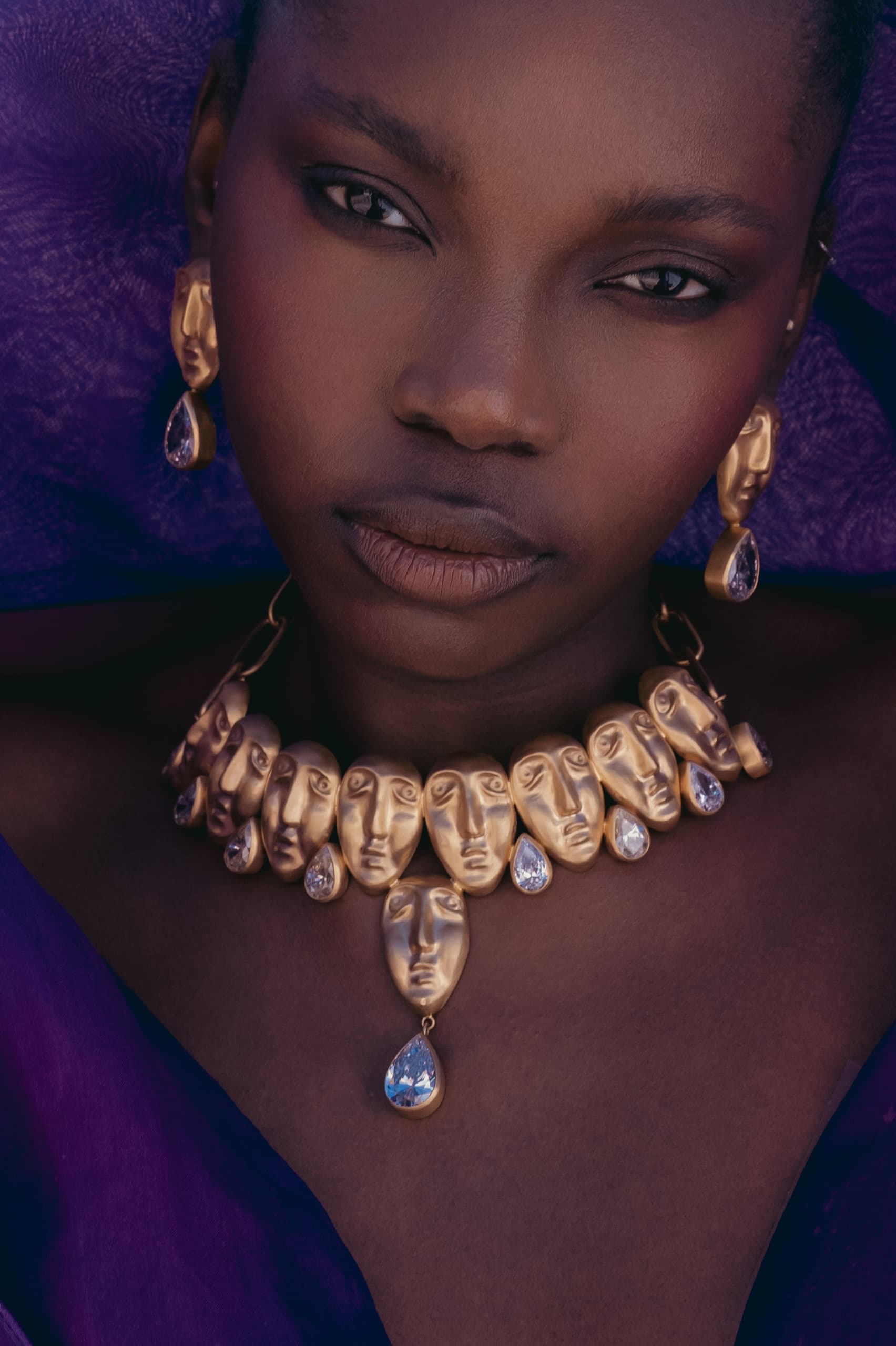 Model wearing artistic face jewelry with gold earrings and necklace, purple attire.