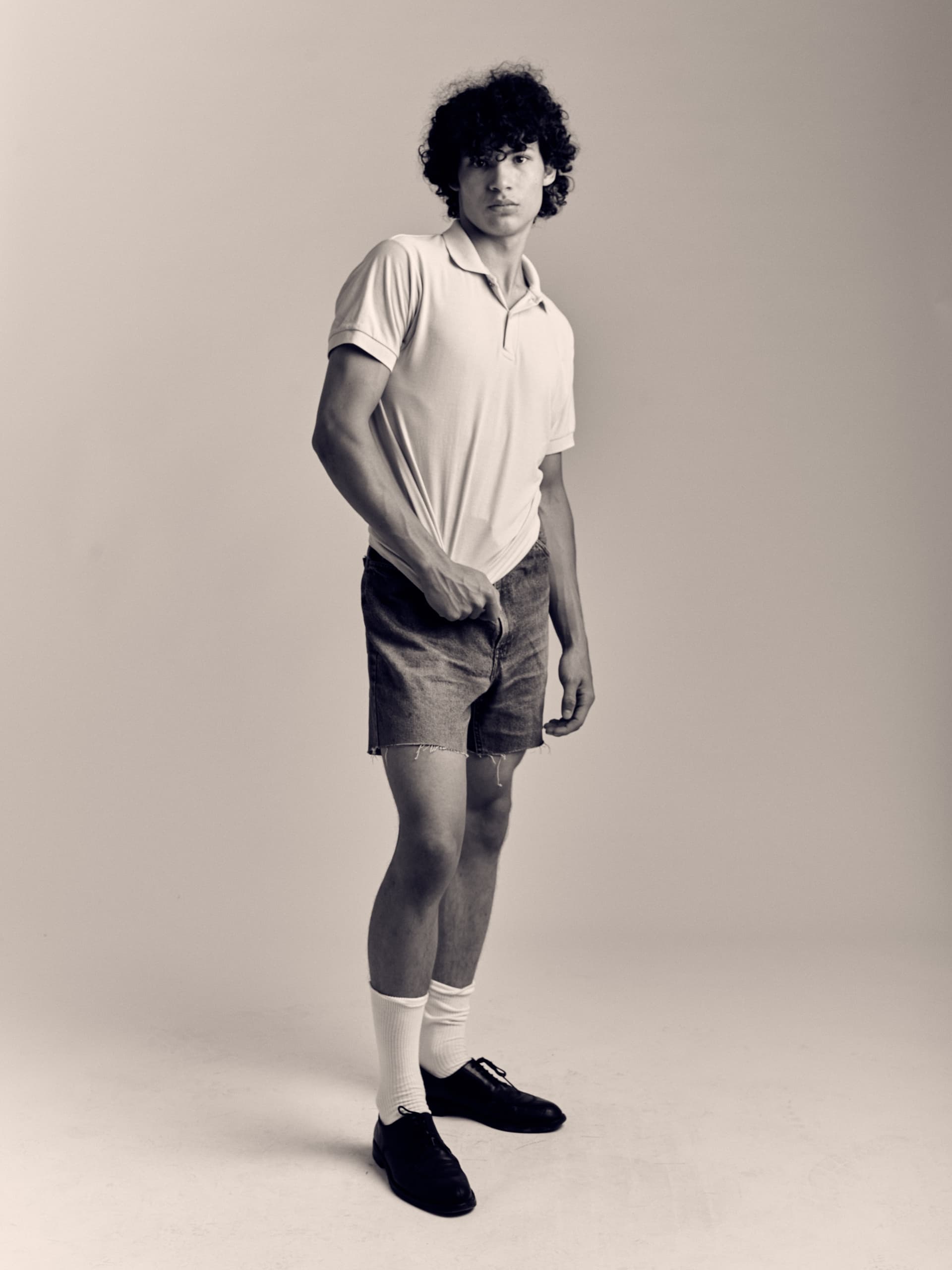 Man in casual retro outfit, curly hair, posing in a monochrome studio setting.