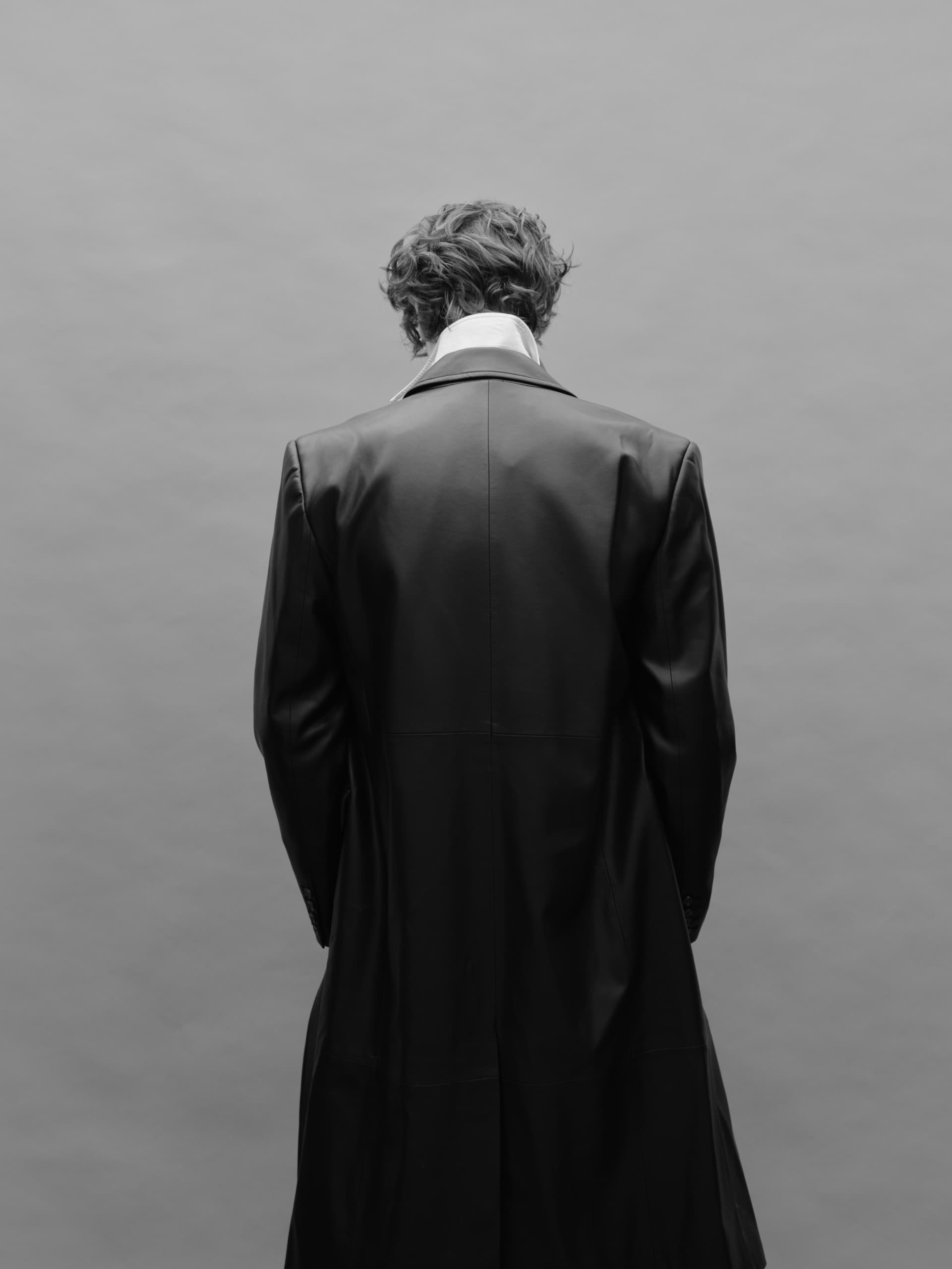 Back view of a person in a long coat, facing a plain grey background.