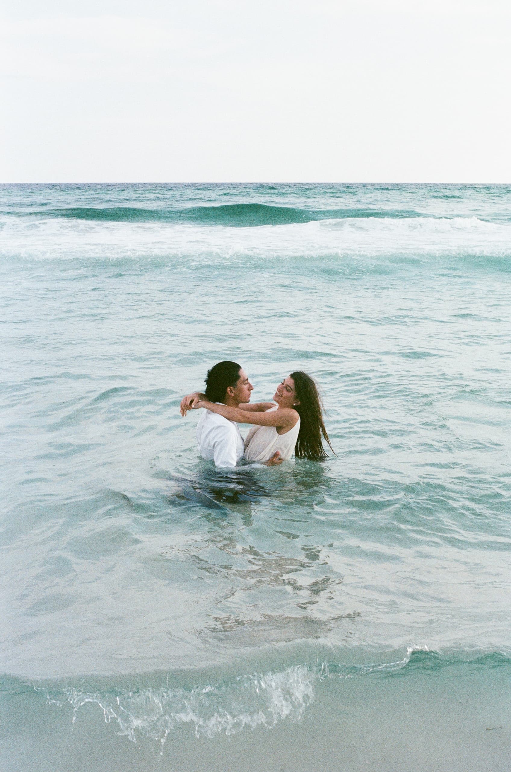 Couple embracing in ocean waves, romantic beach scene, clear sky backdrop.