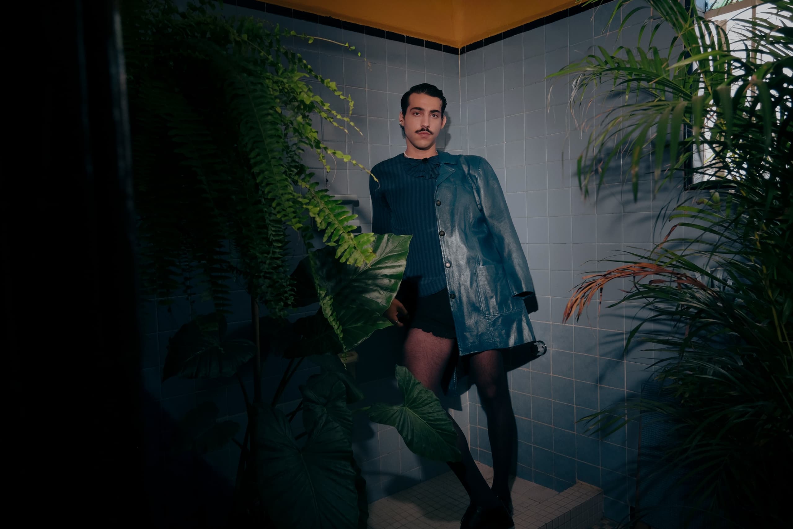Fashionable person in a tiled room with lush green plants and a blue coat.