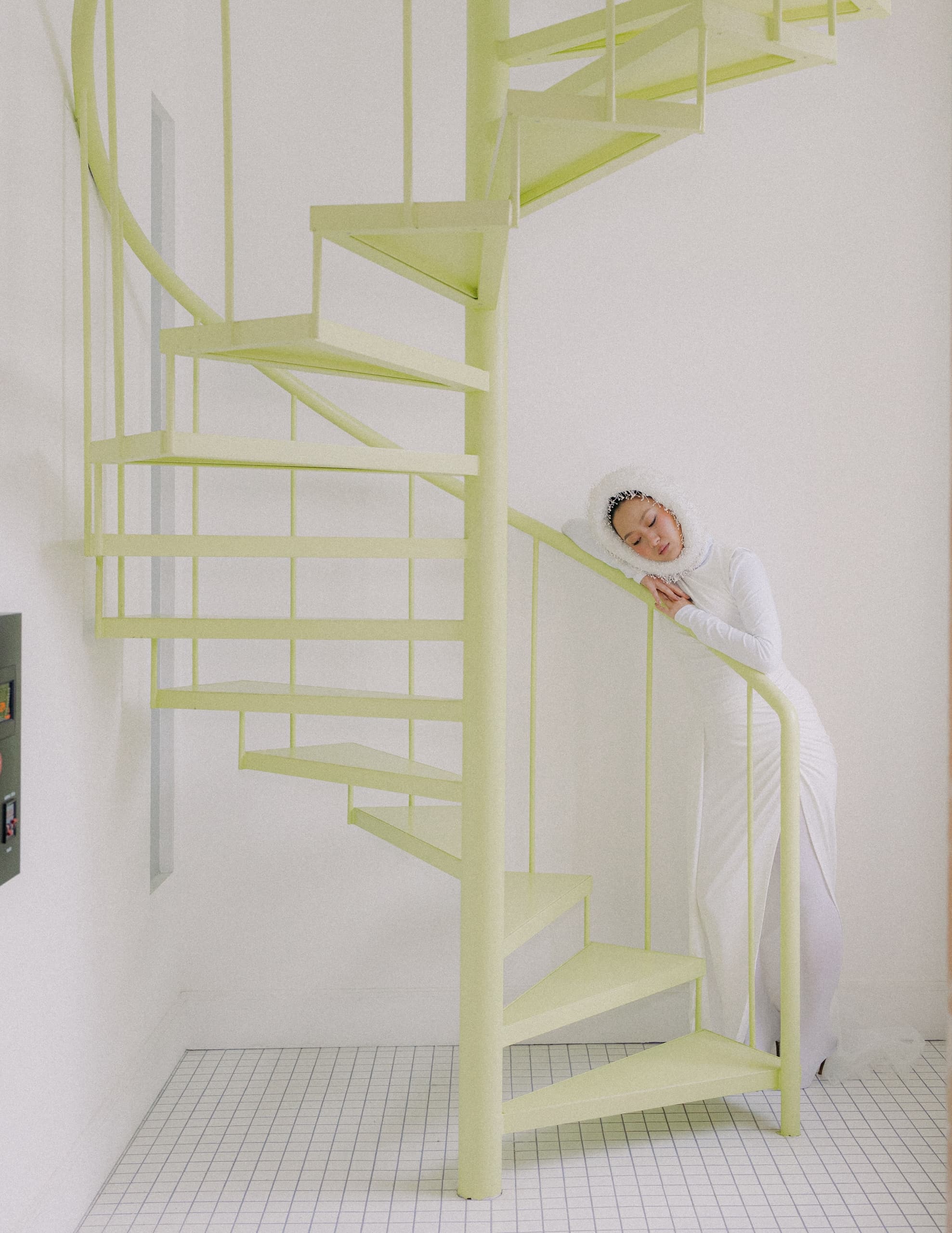 Person in white outfit beside pastel spiral staircase in minimalist setting.