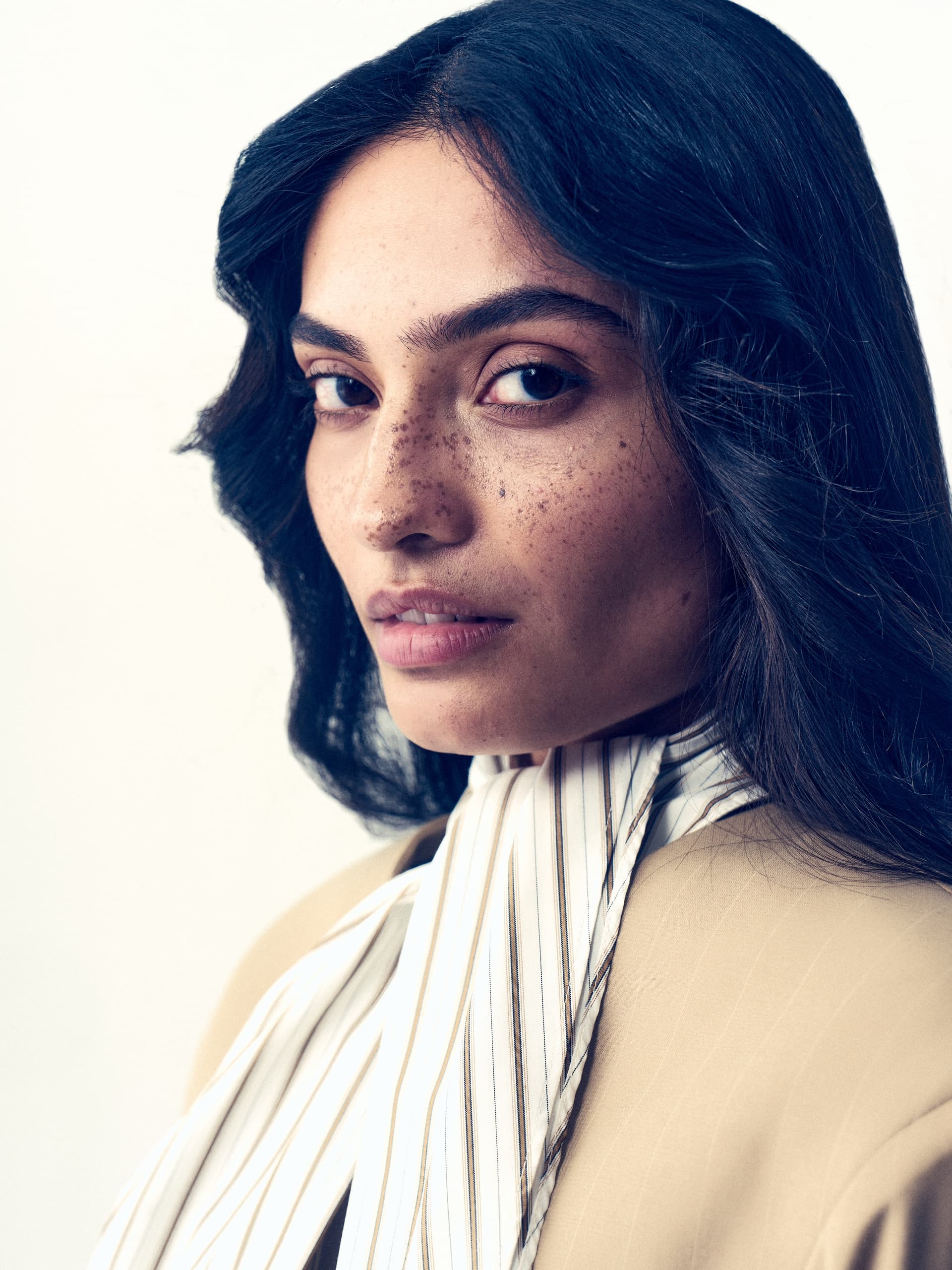 Close-up of a woman with freckles wearing a beige blazer and striped scarf.