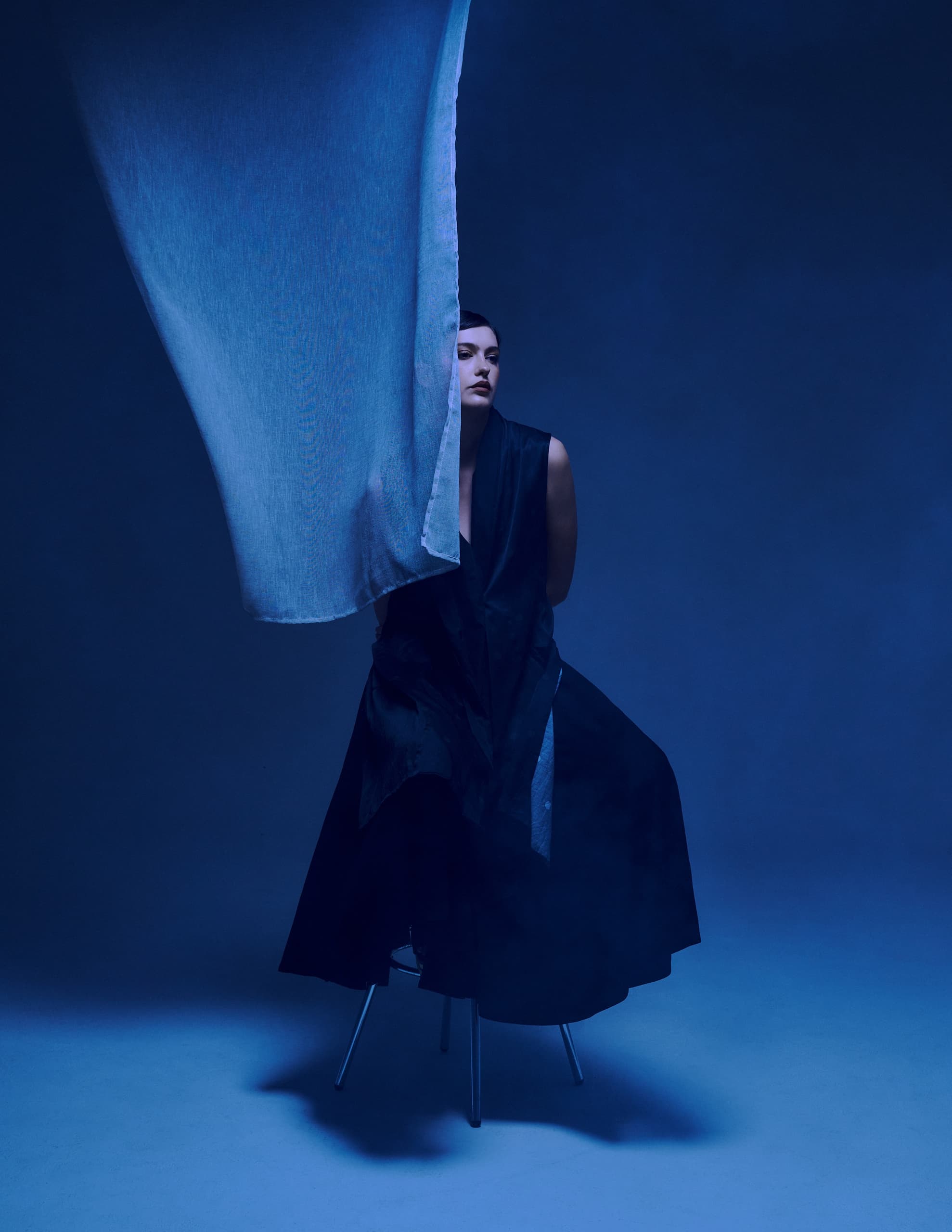 Woman in dark dress partially obscured by flowing blue fabric, seated on a stool.