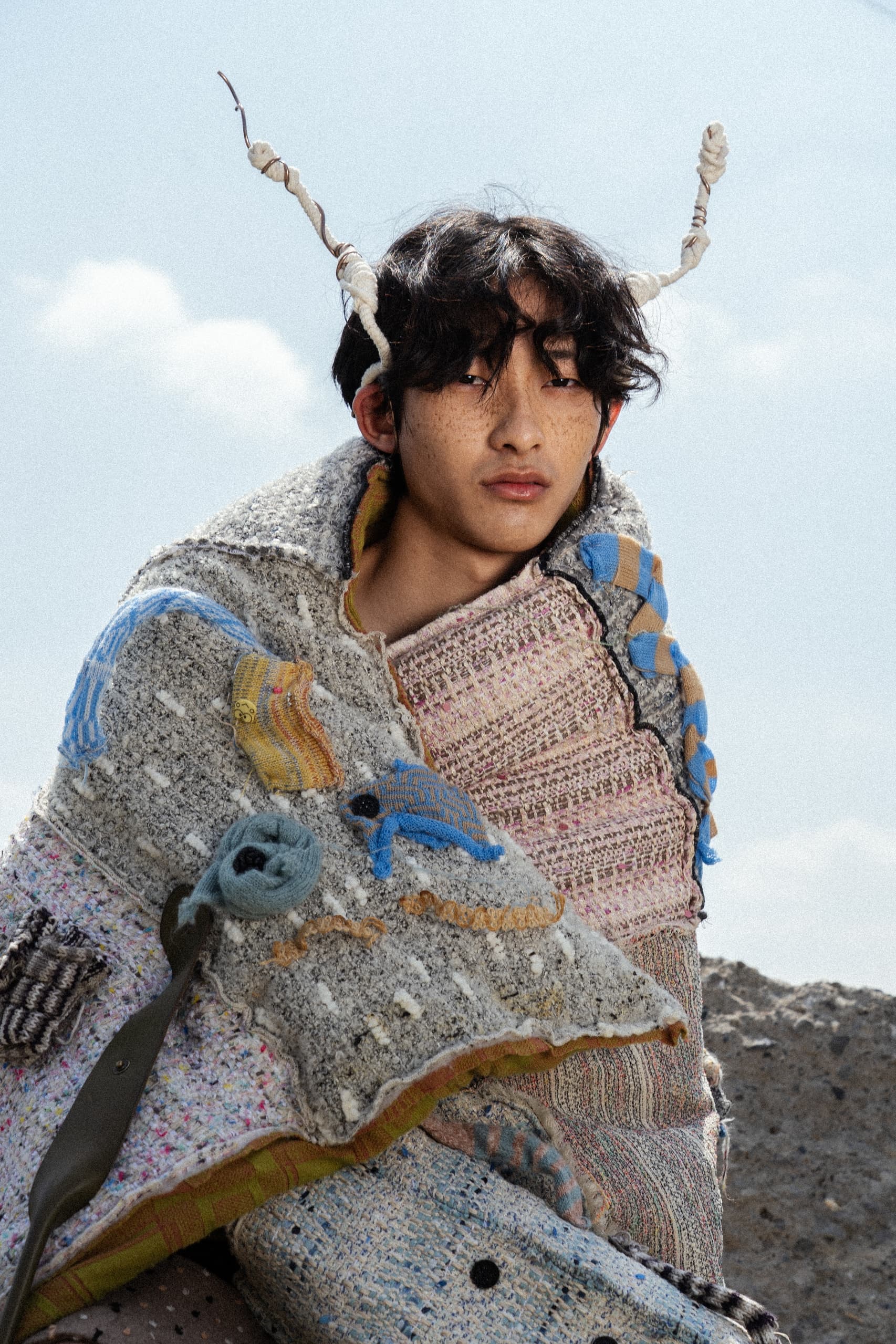 Person wears eclectic patchwork attire with twisted headpiece under a clear sky.