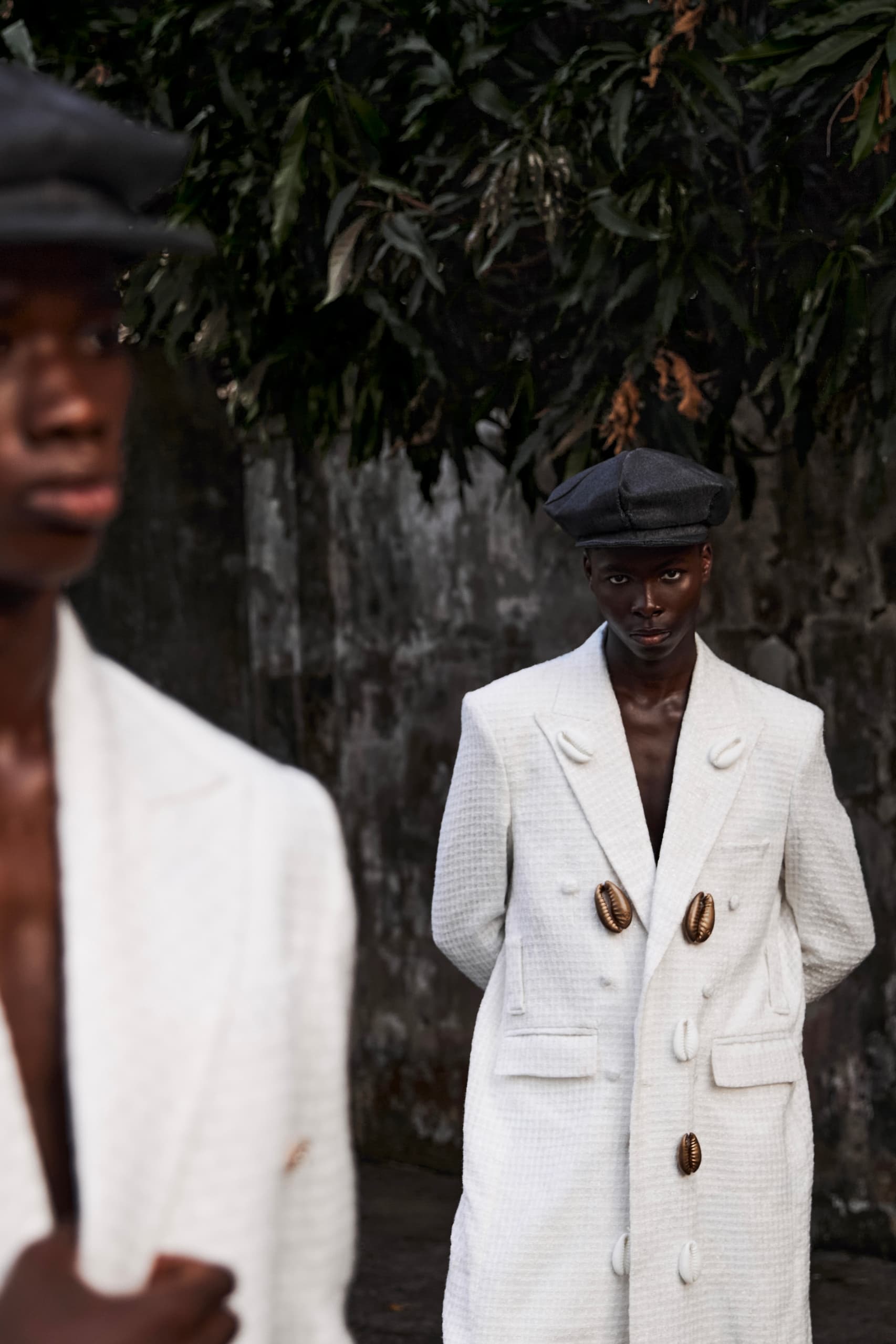 Stylish models in white coats and hats against a leafy background.