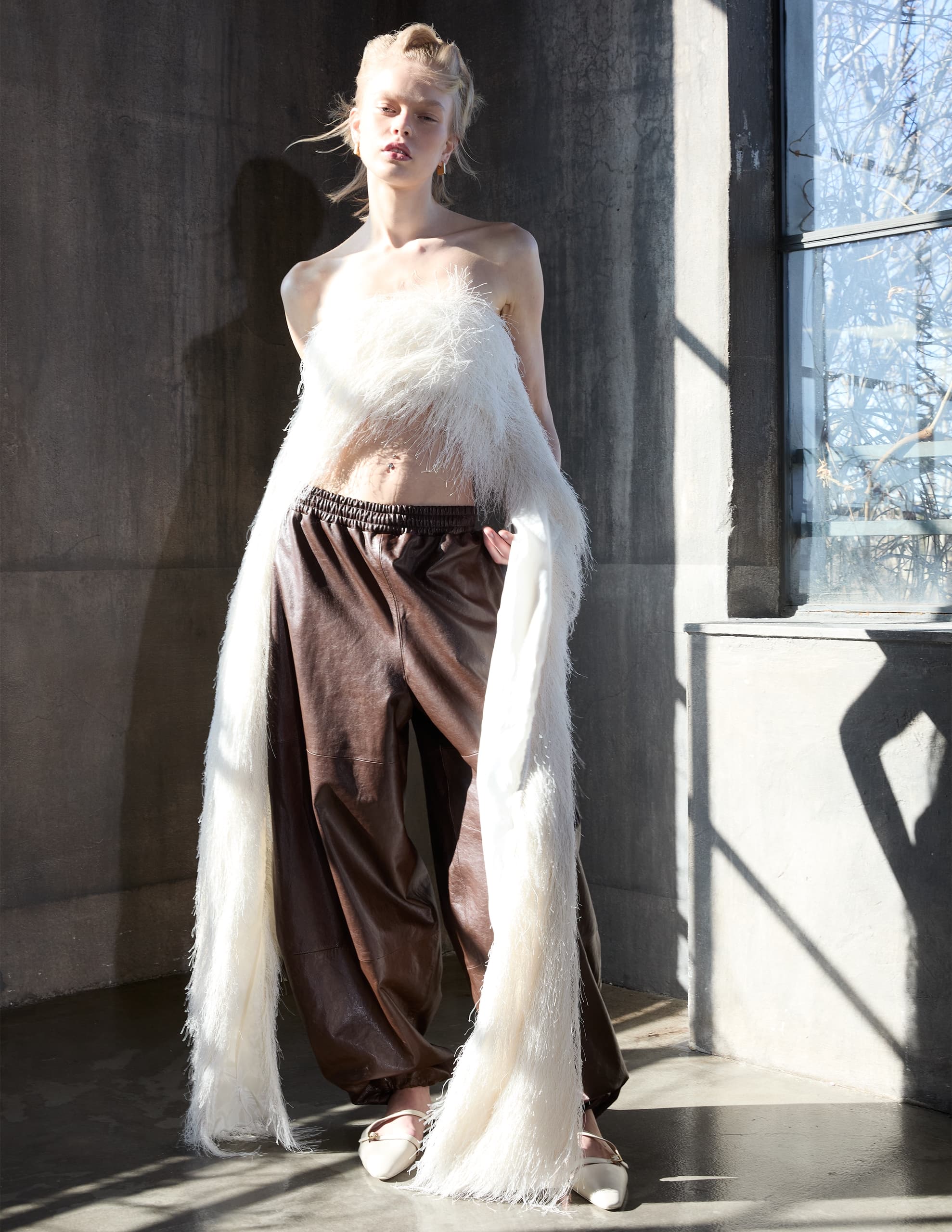 Stylish model in fashionable fur top and leather pants by a sunlit window.