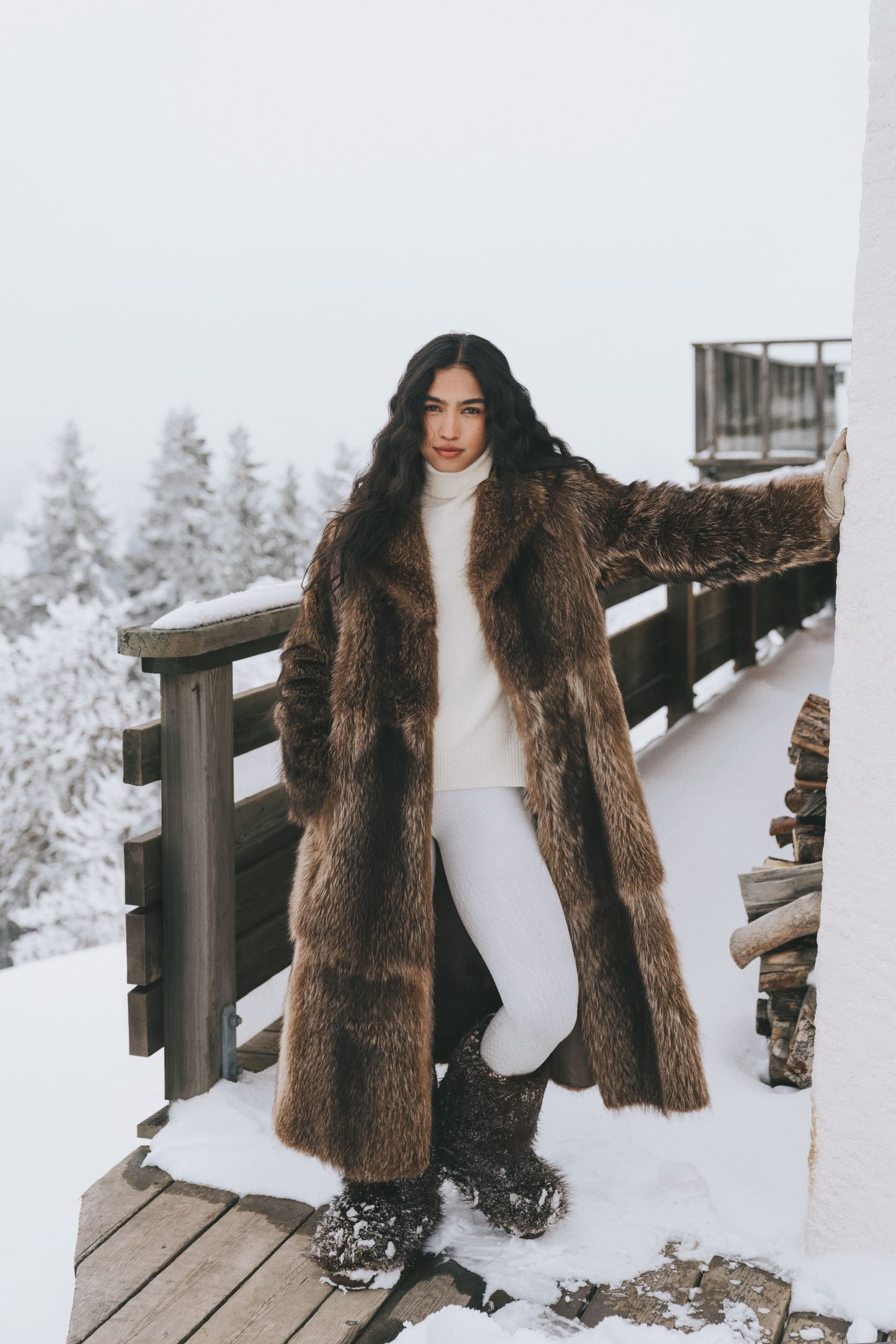 Woman in fur coat stands on snowy deck with winter forest backdrop.