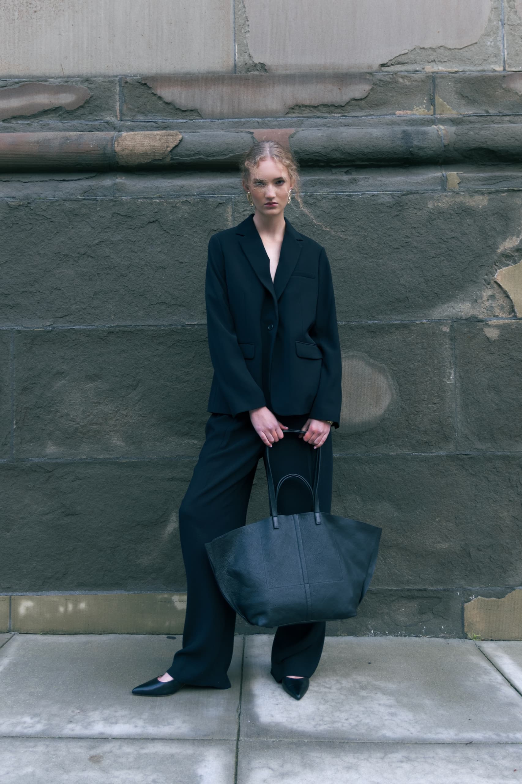 Fashionable woman in black suit holding a large tote bag against a stone wall.