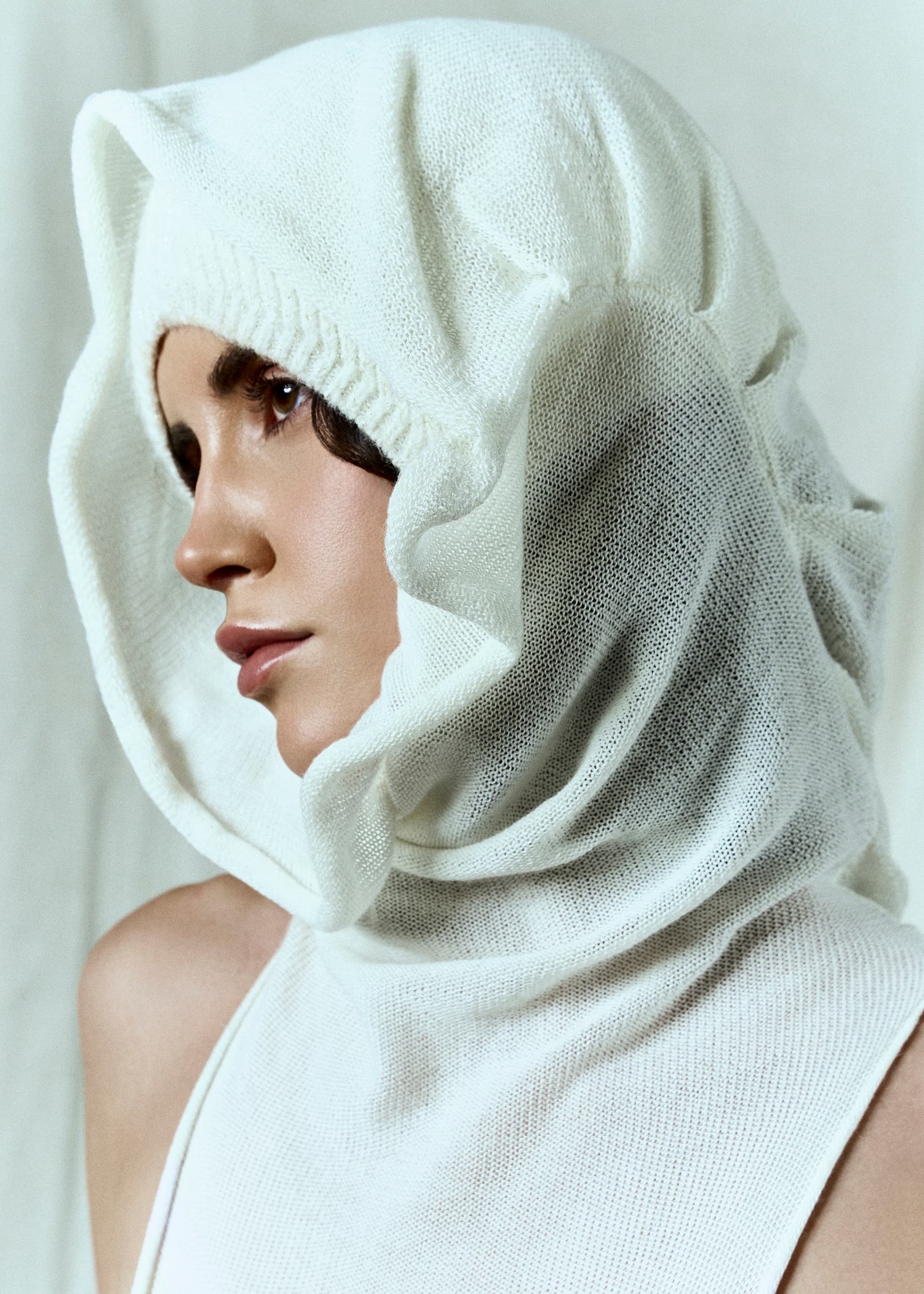 Woman in white knitted hooded garment, side profile, minimalist fashion photography.