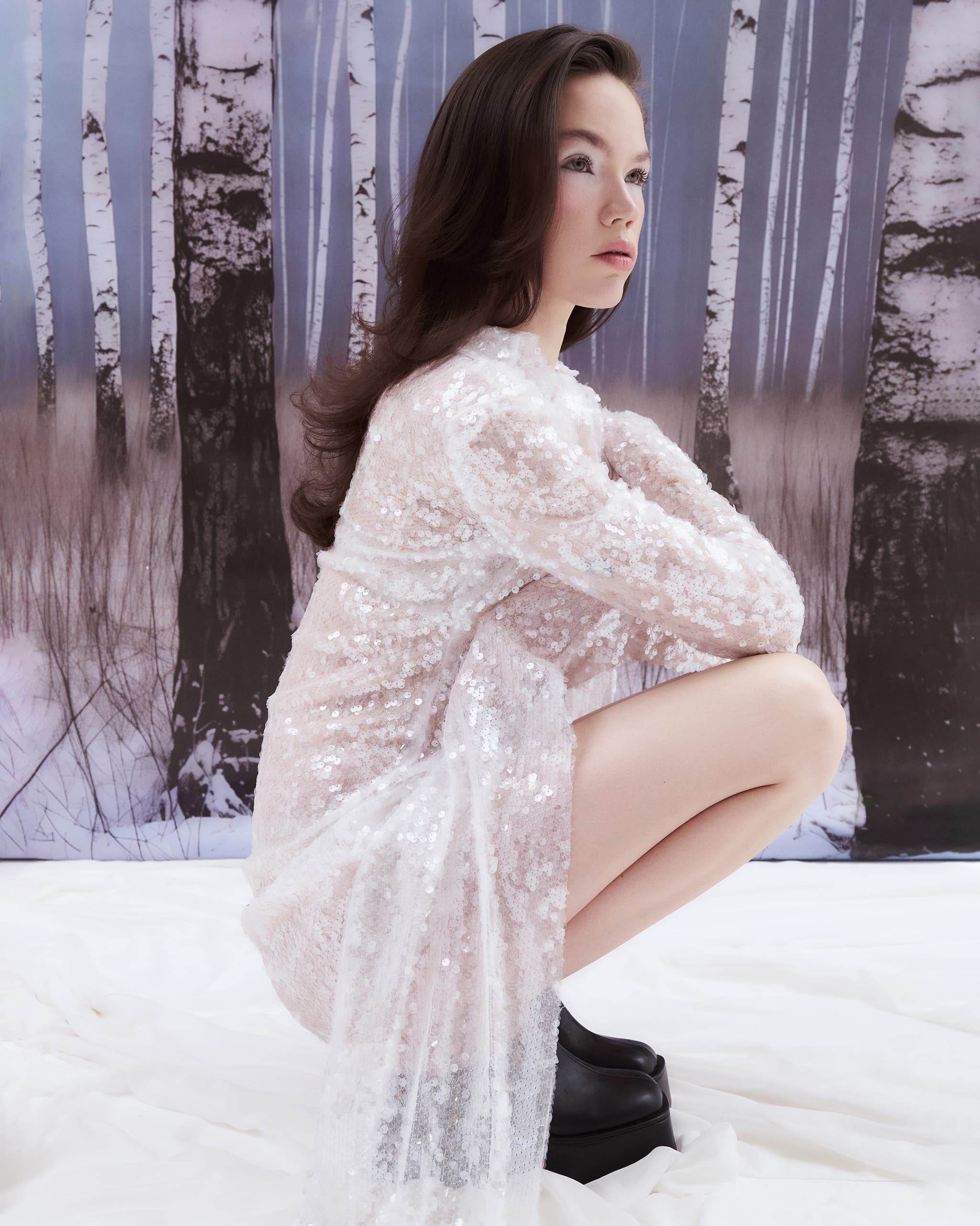 Woman in sequin dress posing in front of a snowy birch forest backdrop.