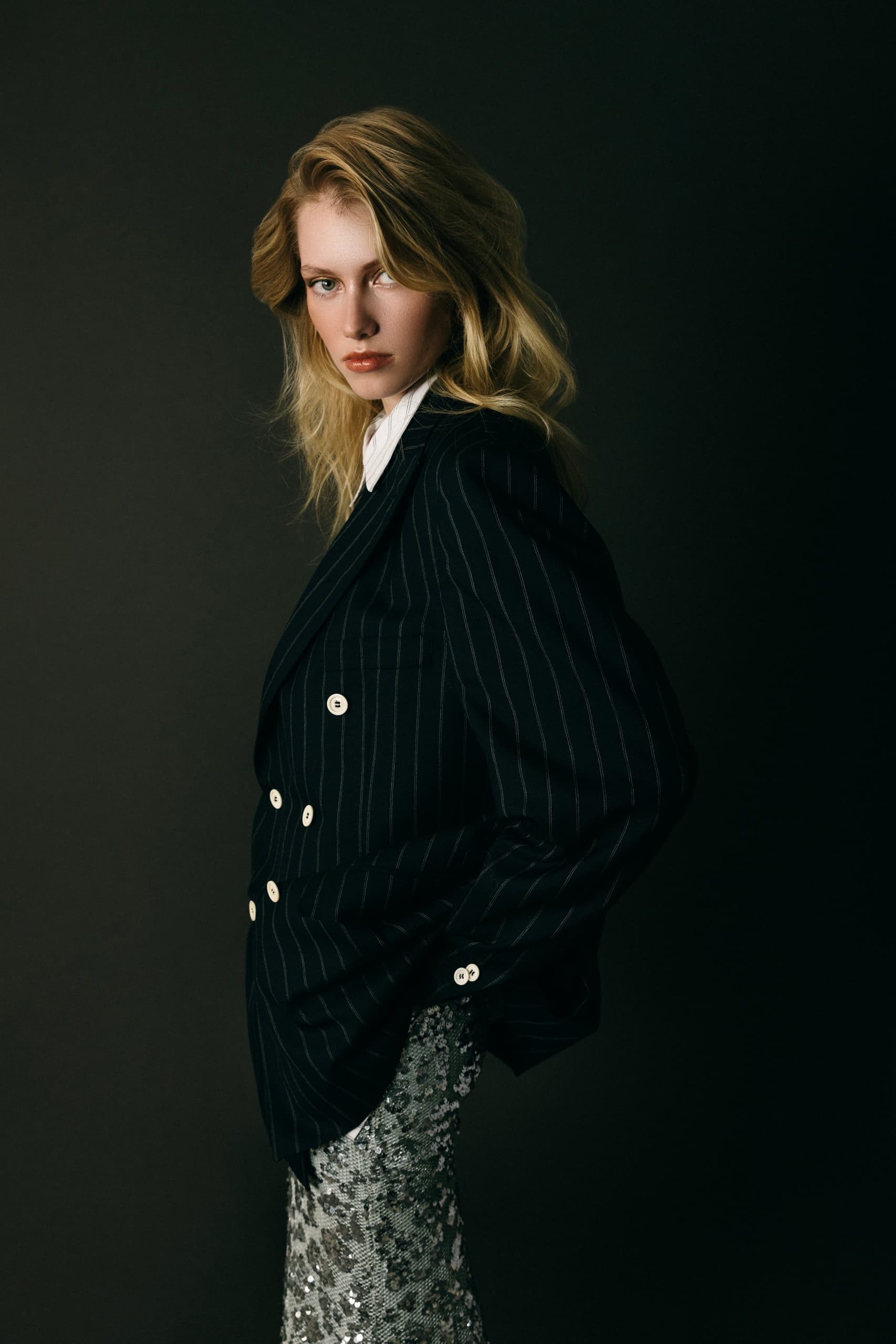 Stylish woman in black pinstripe suit with sequined skirt, dark backdrop.