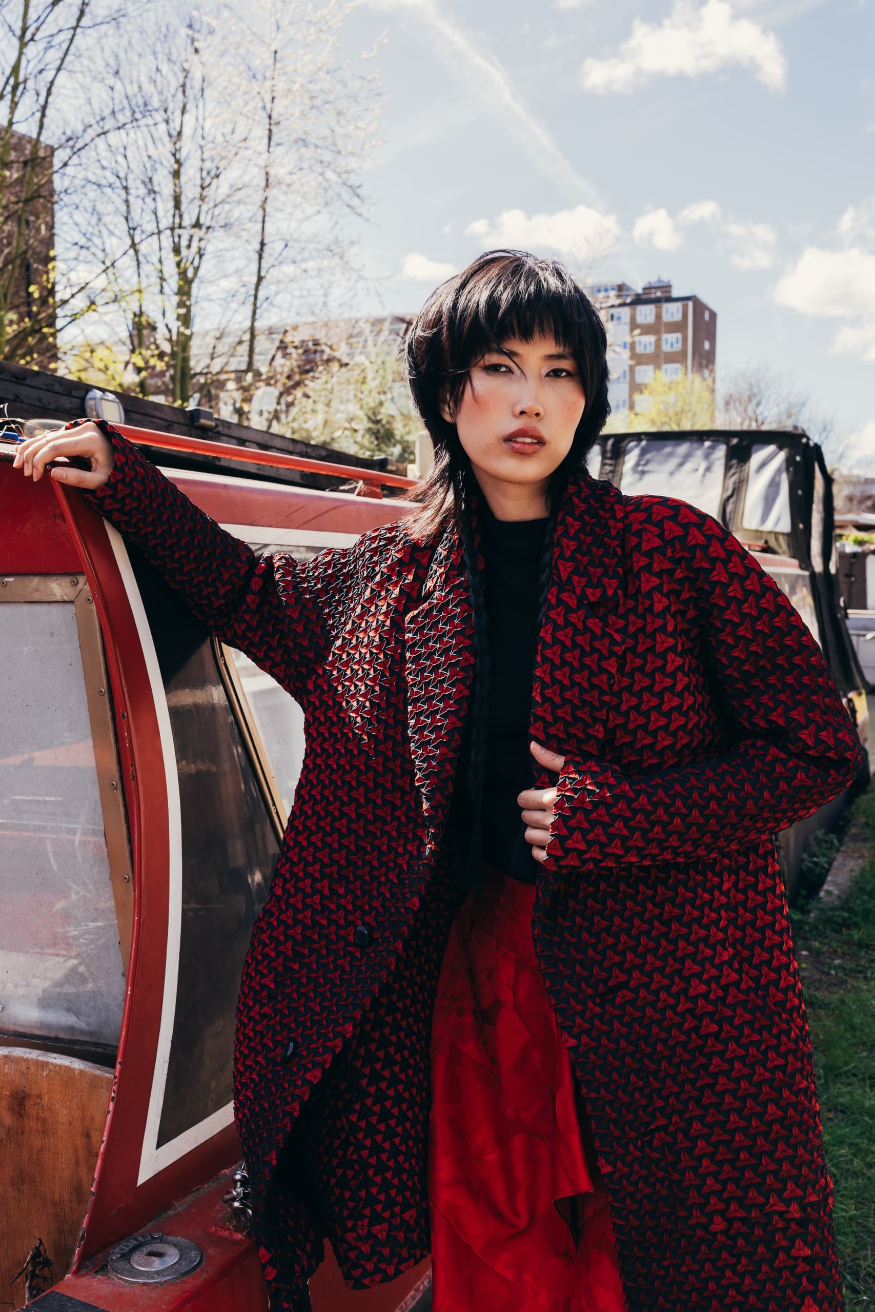 Fashionable person in red geometric coat leaning against a boat outdoors.