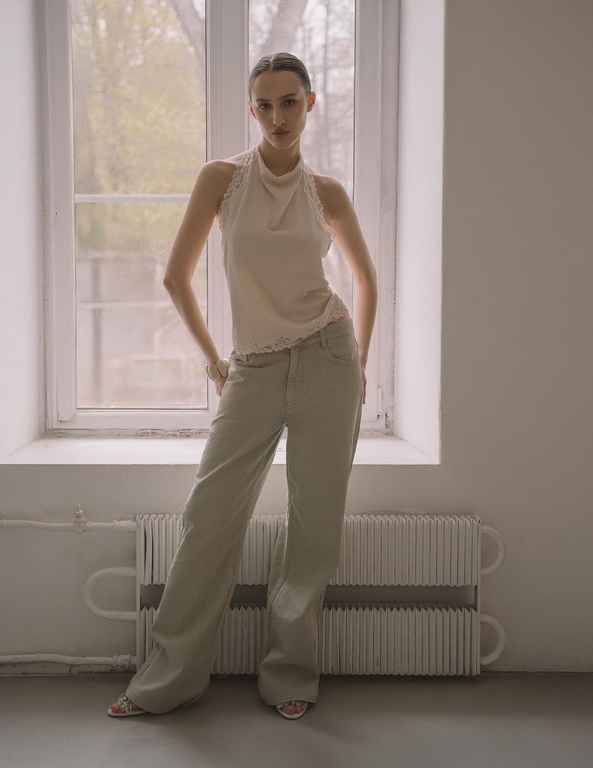 Woman in beige top and green pants standing by a window, minimalist fashion style.