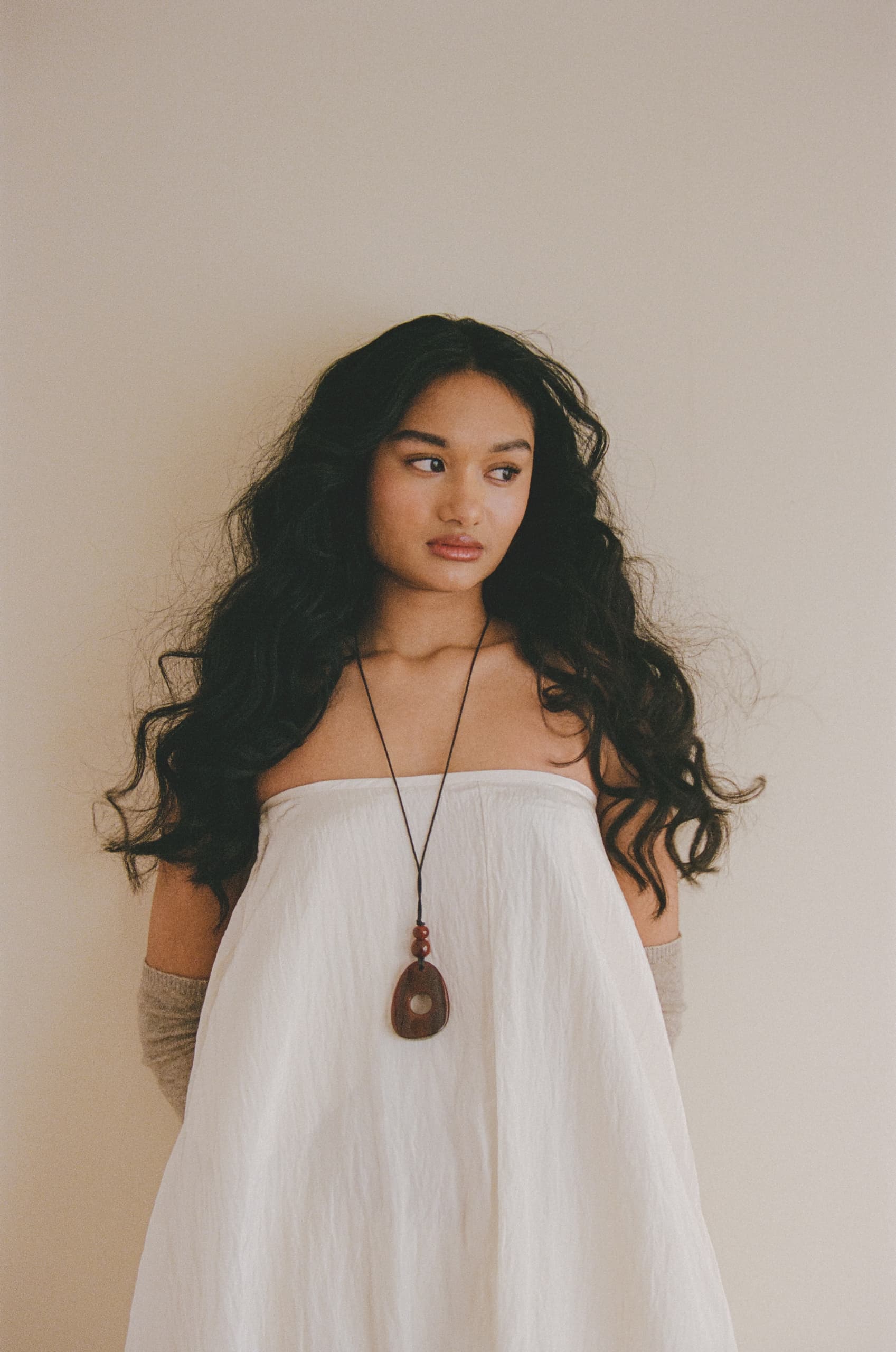 Woman in white dress with long hair and pendant, standing against a neutral background.