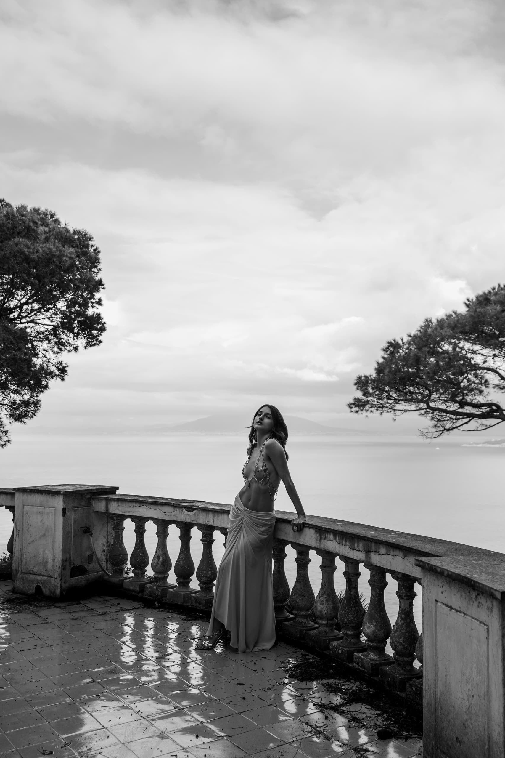 Woman in elegant dress stands on balcony overlooking ocean in black and white.