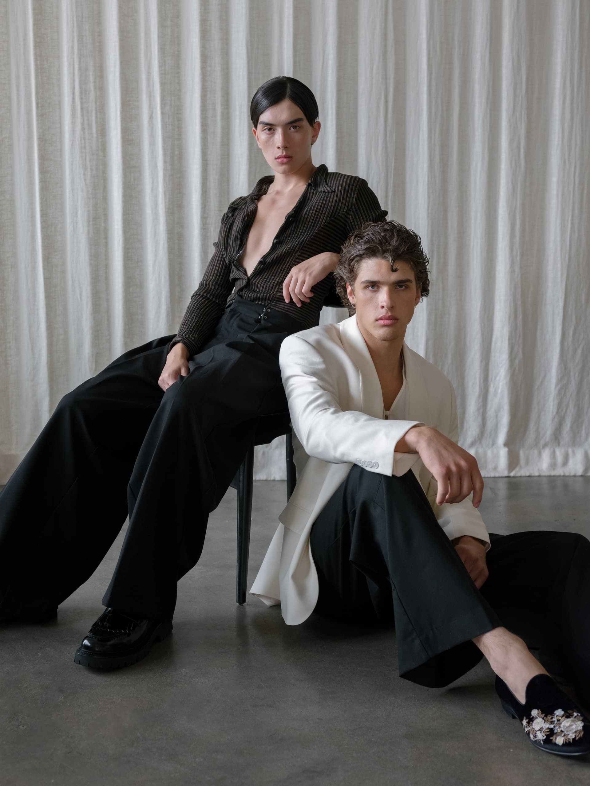 Two models in stylish outfits pose against a curtain backdrop.