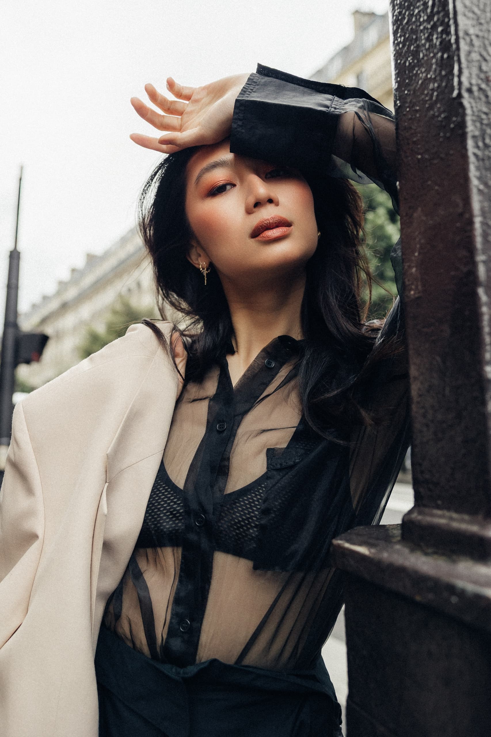 Fashionable woman poses in city street, wearing sheer black blouse and beige jacket.