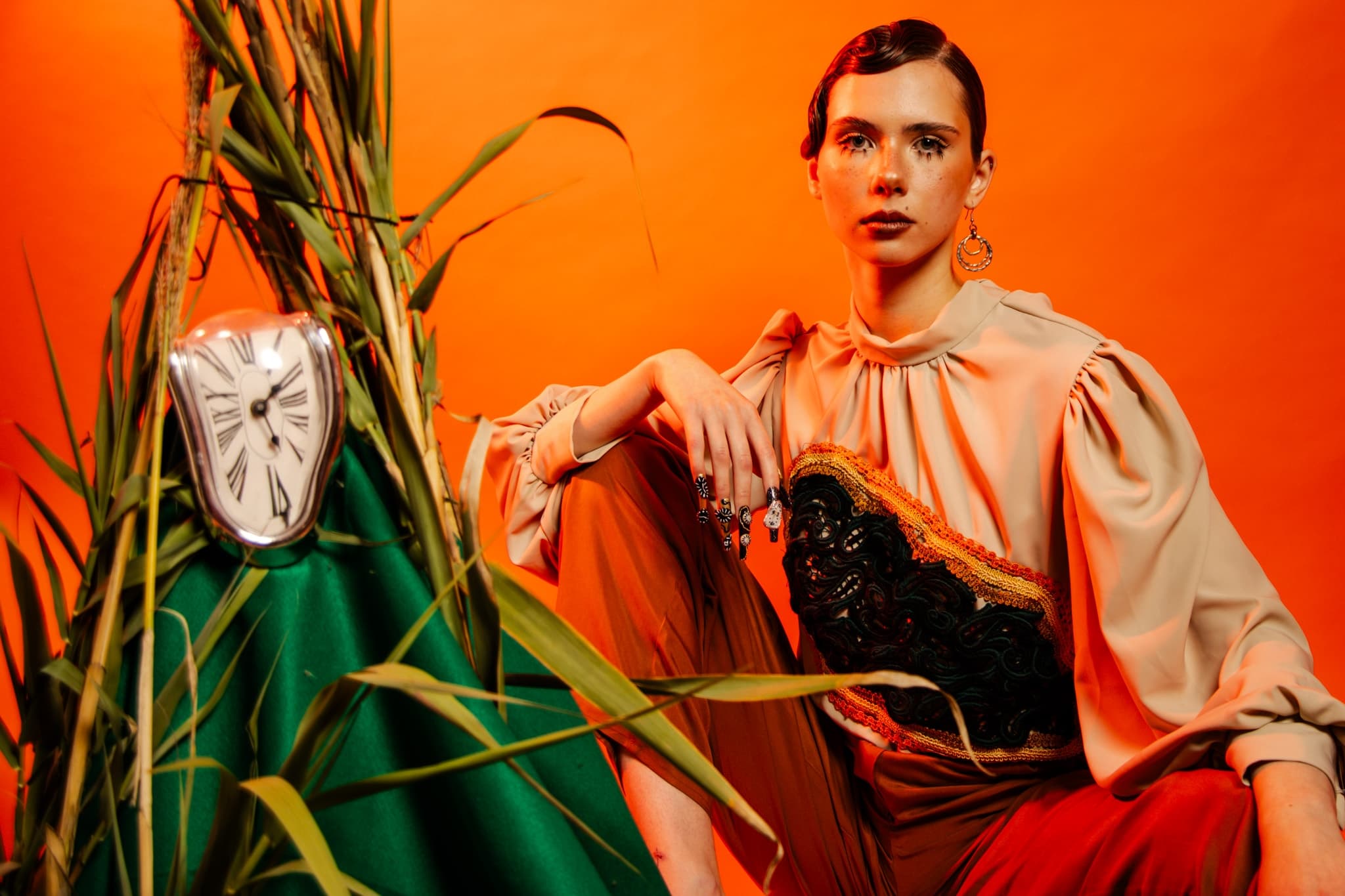 Fashionable woman poses with surreal clock and leaves on orange background.