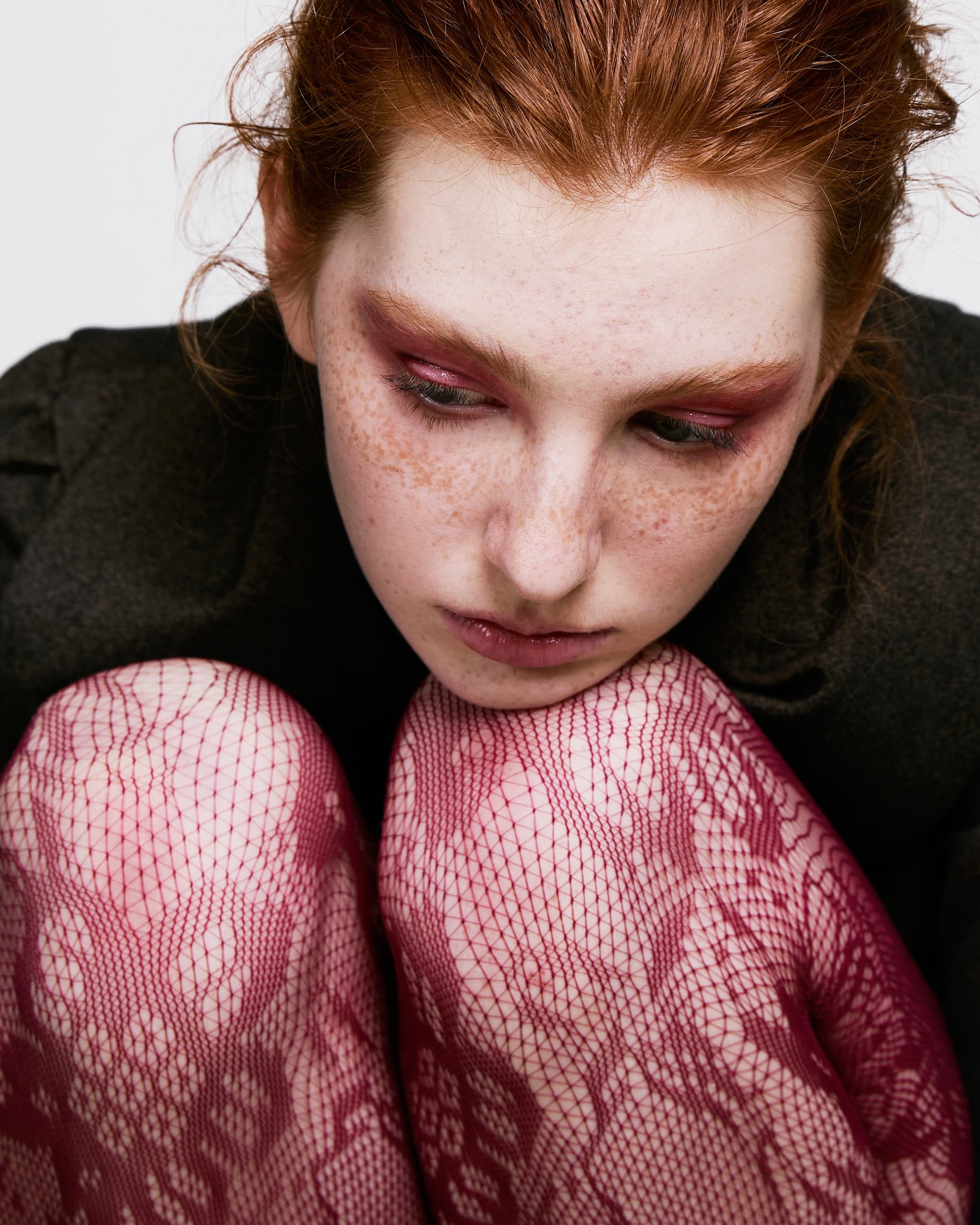 Red-haired woman with pink lace tights and artistic makeup looks pensive.
