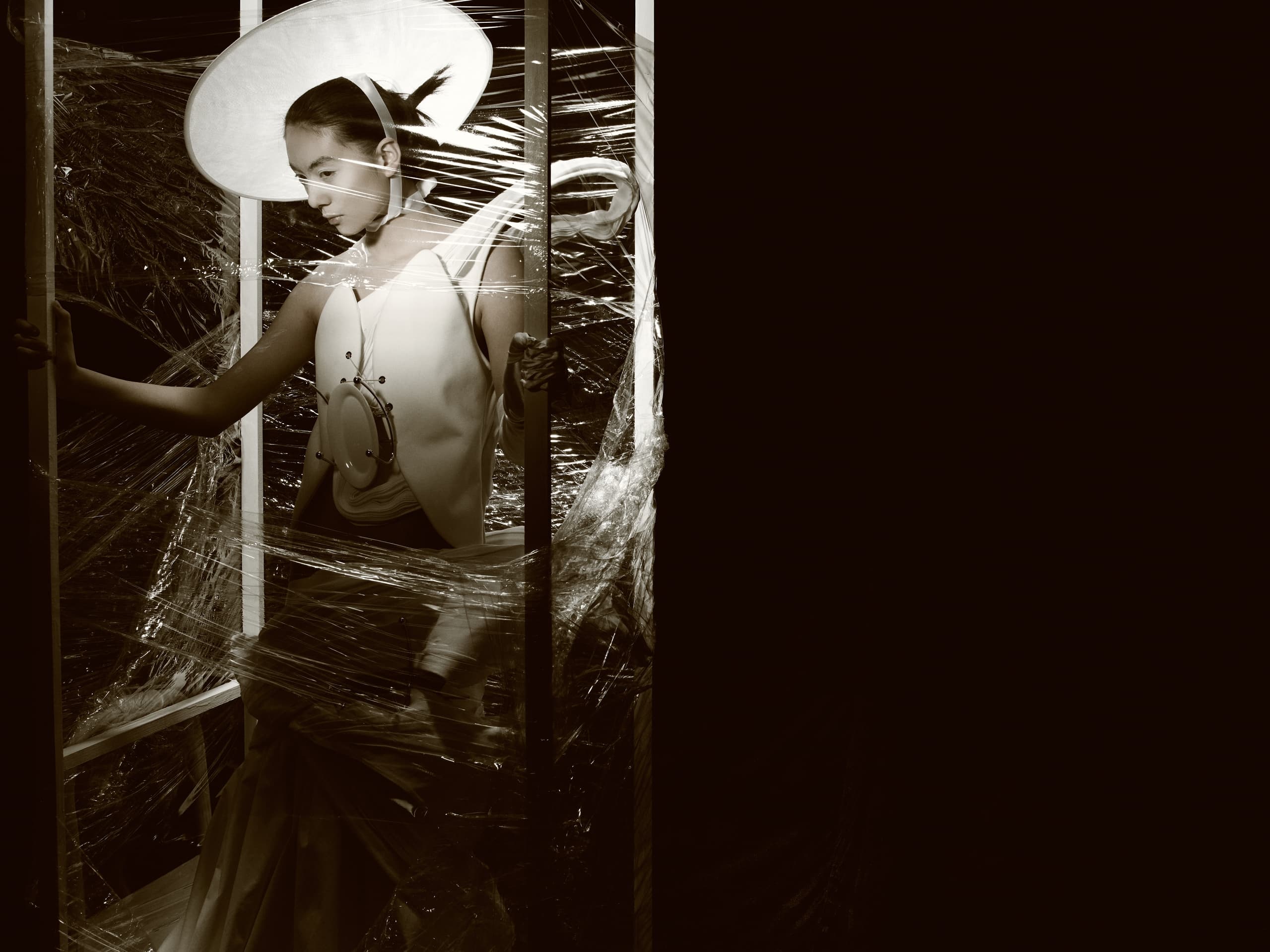 Fashion model in avant-garde outfit and hat, wrapped in plastic, sepia tone.