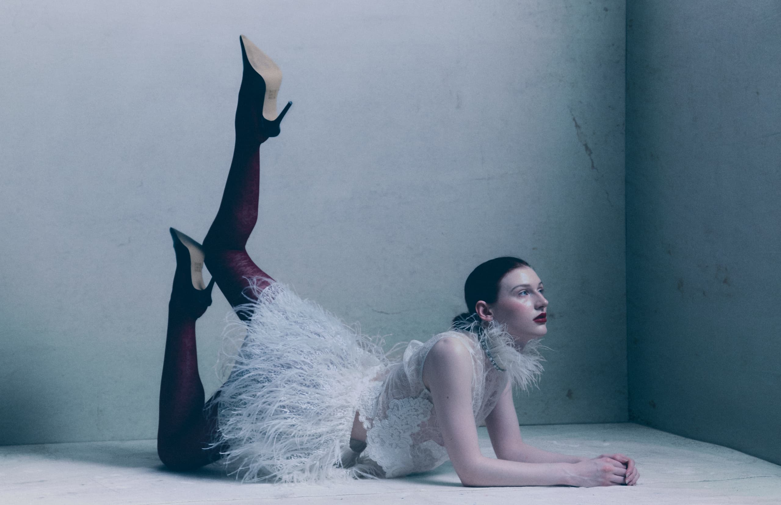 Elegant woman in feather outfit and heels posing in minimalist corner.