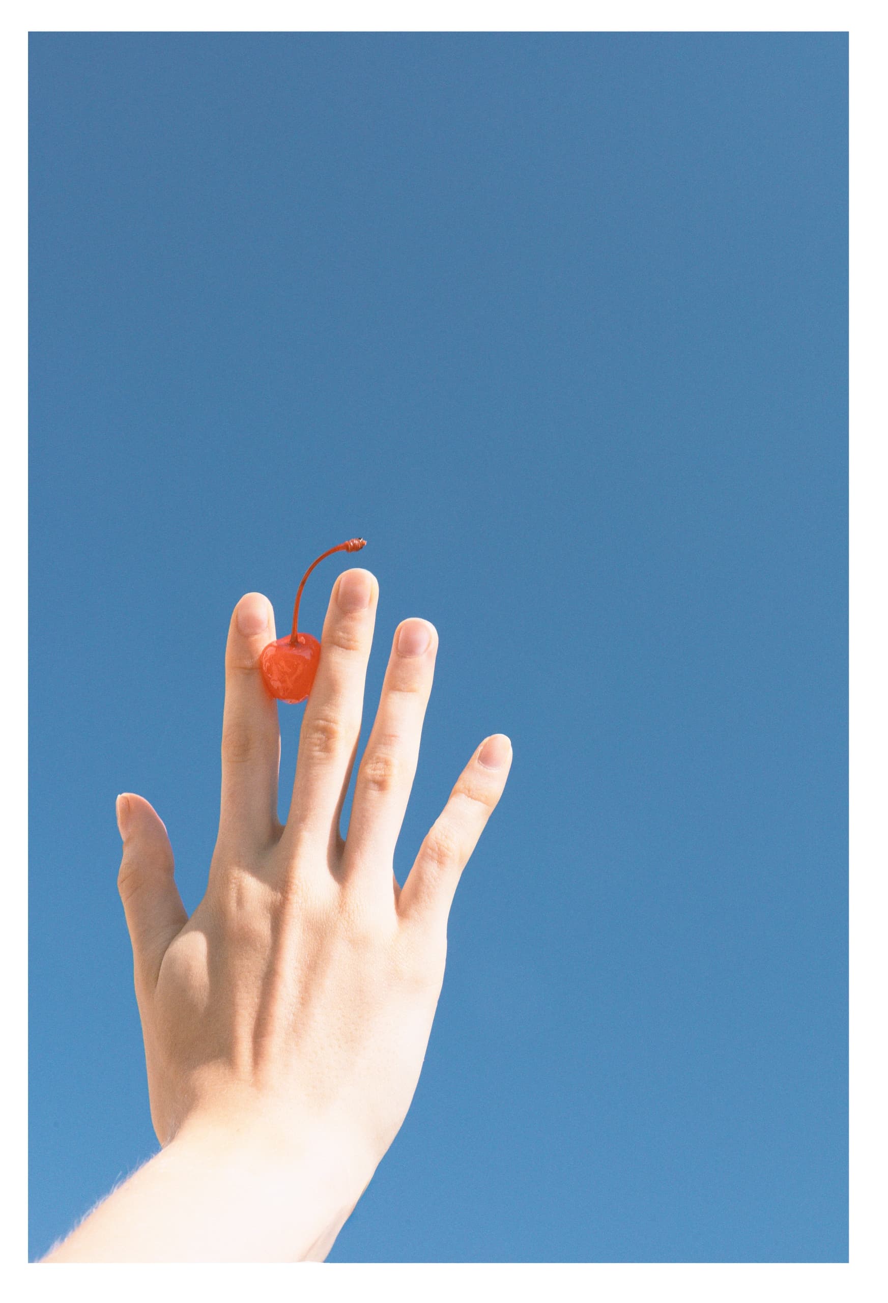 Hand balancing a cherry under a clear blue sky.