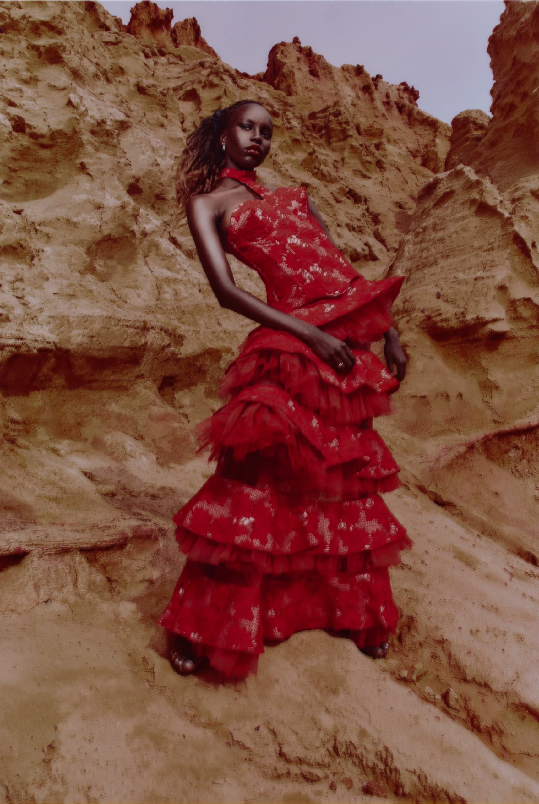 Model poses in layered red dress against rocky desert backdrop.