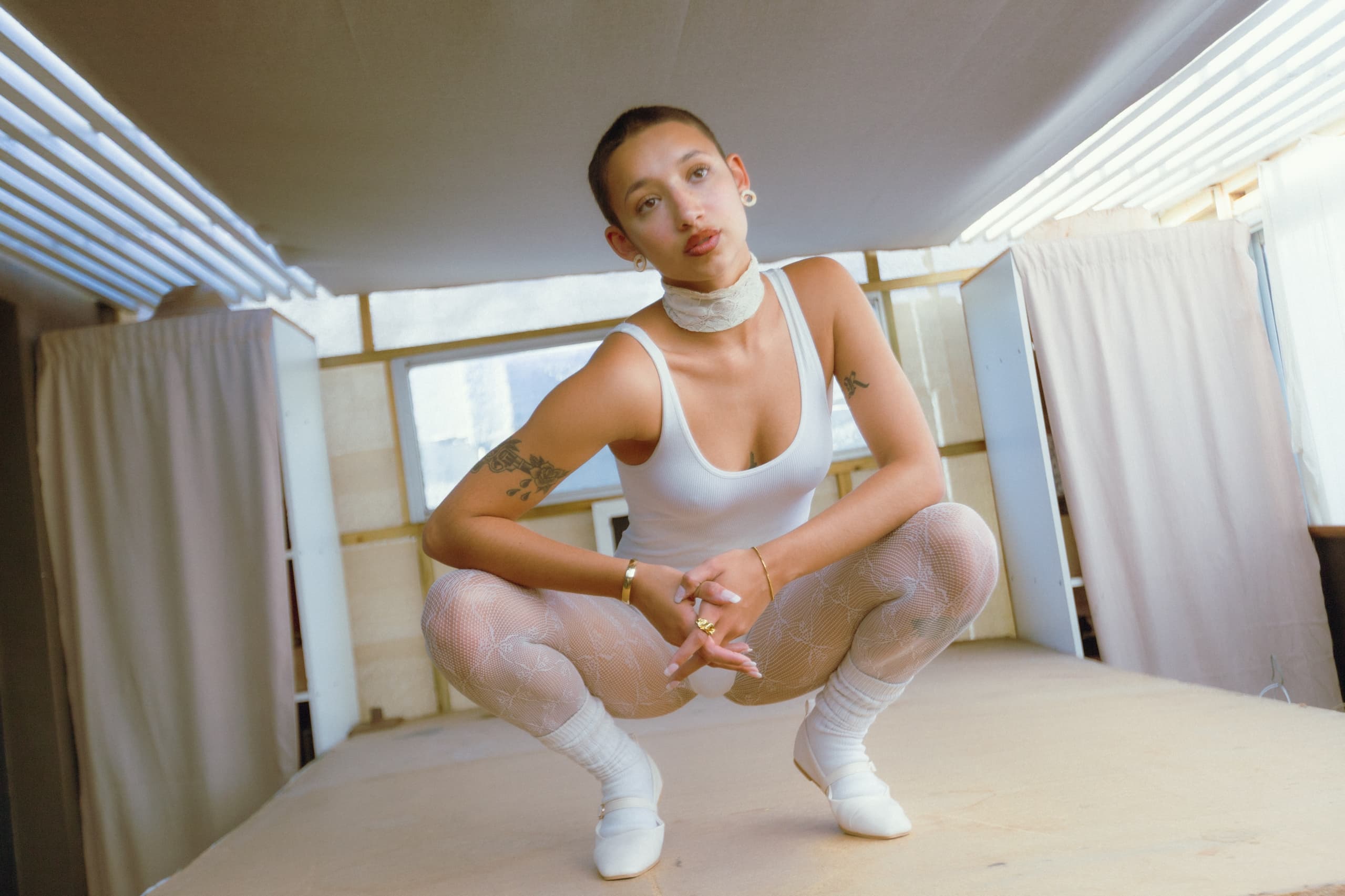 Person in stylish white outfit and tattoos, crouching indoors. Fashionable and modern look.