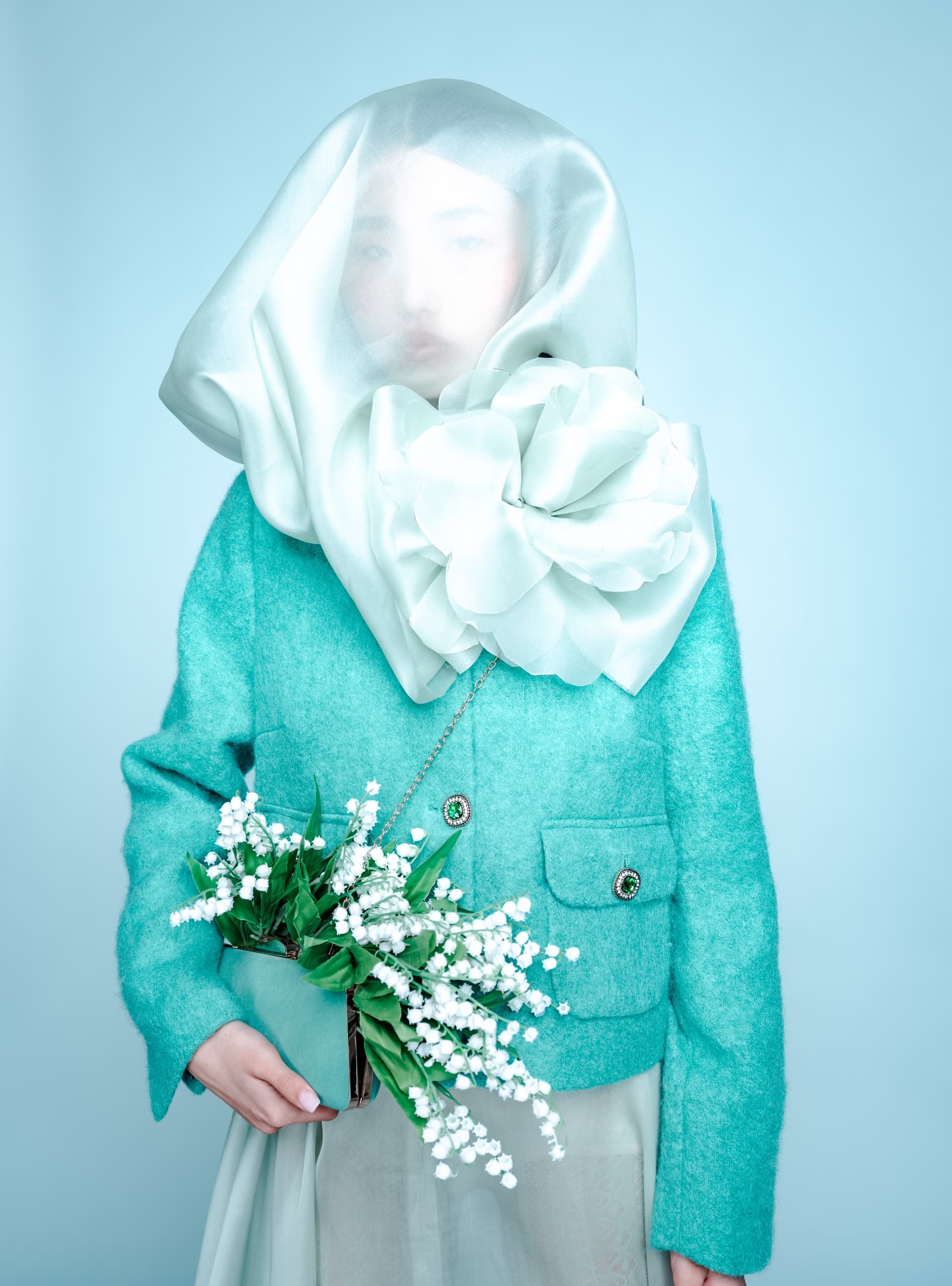 Woman in teal with sheer veil and flower accessory, holding lily of the valley bouquet.