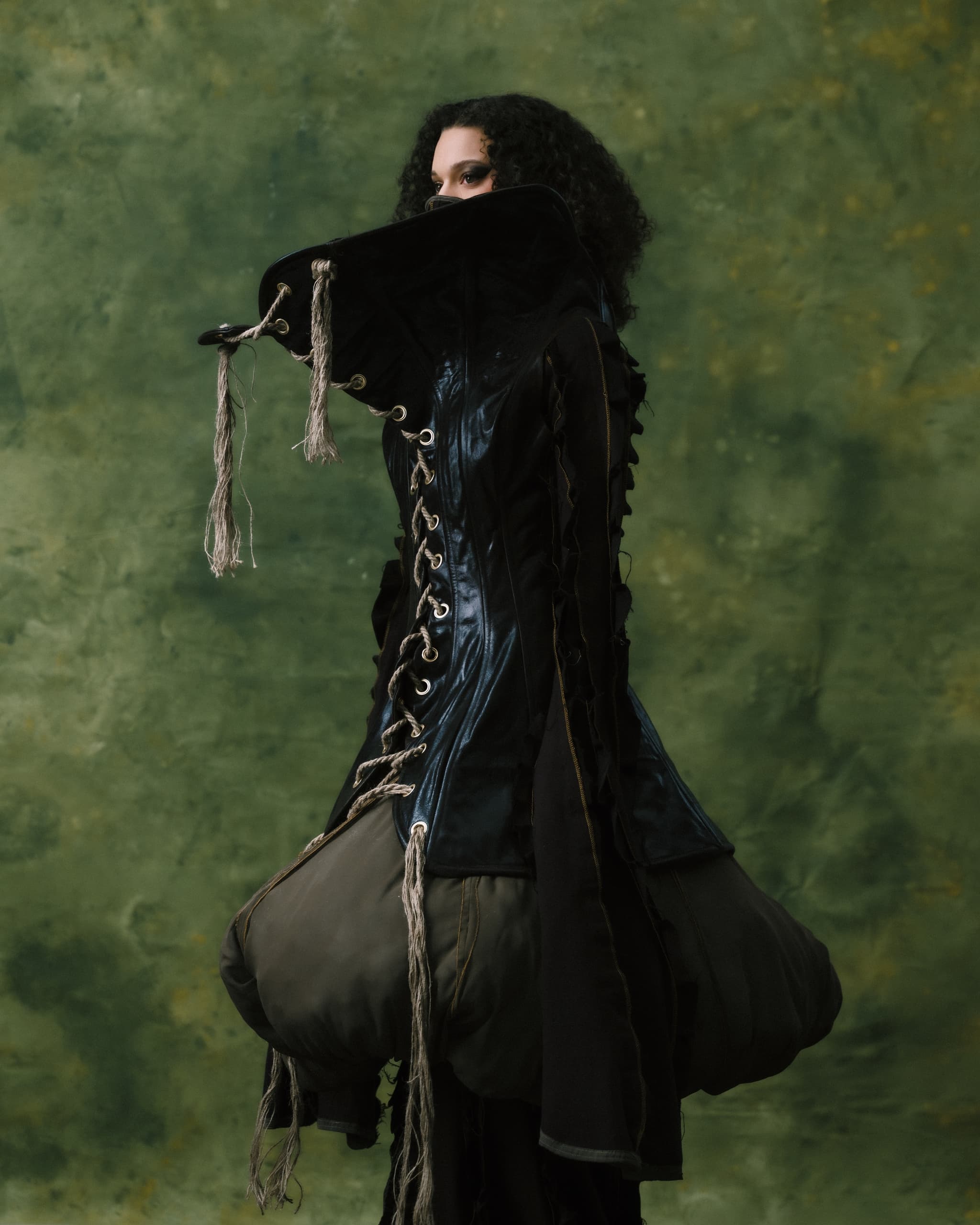 Gothic fashion model in a dramatic, dark outfit against a textured green background.