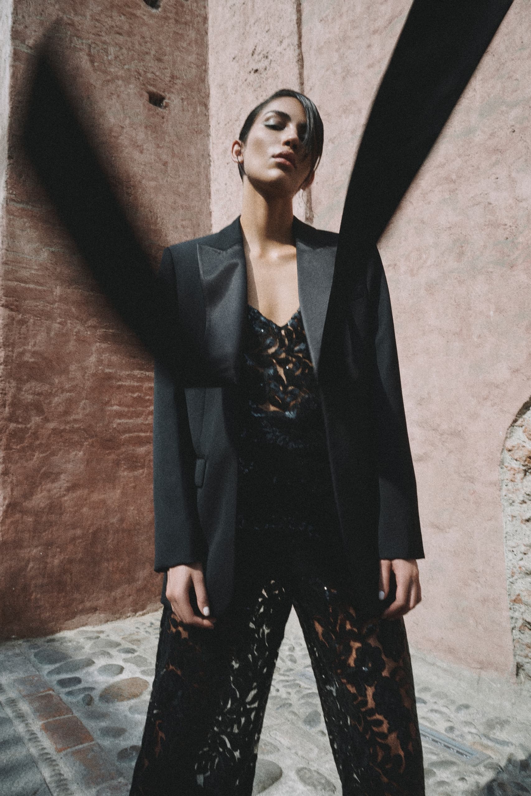 Stylish person in black lace outfit and blazer against rustic wall background.