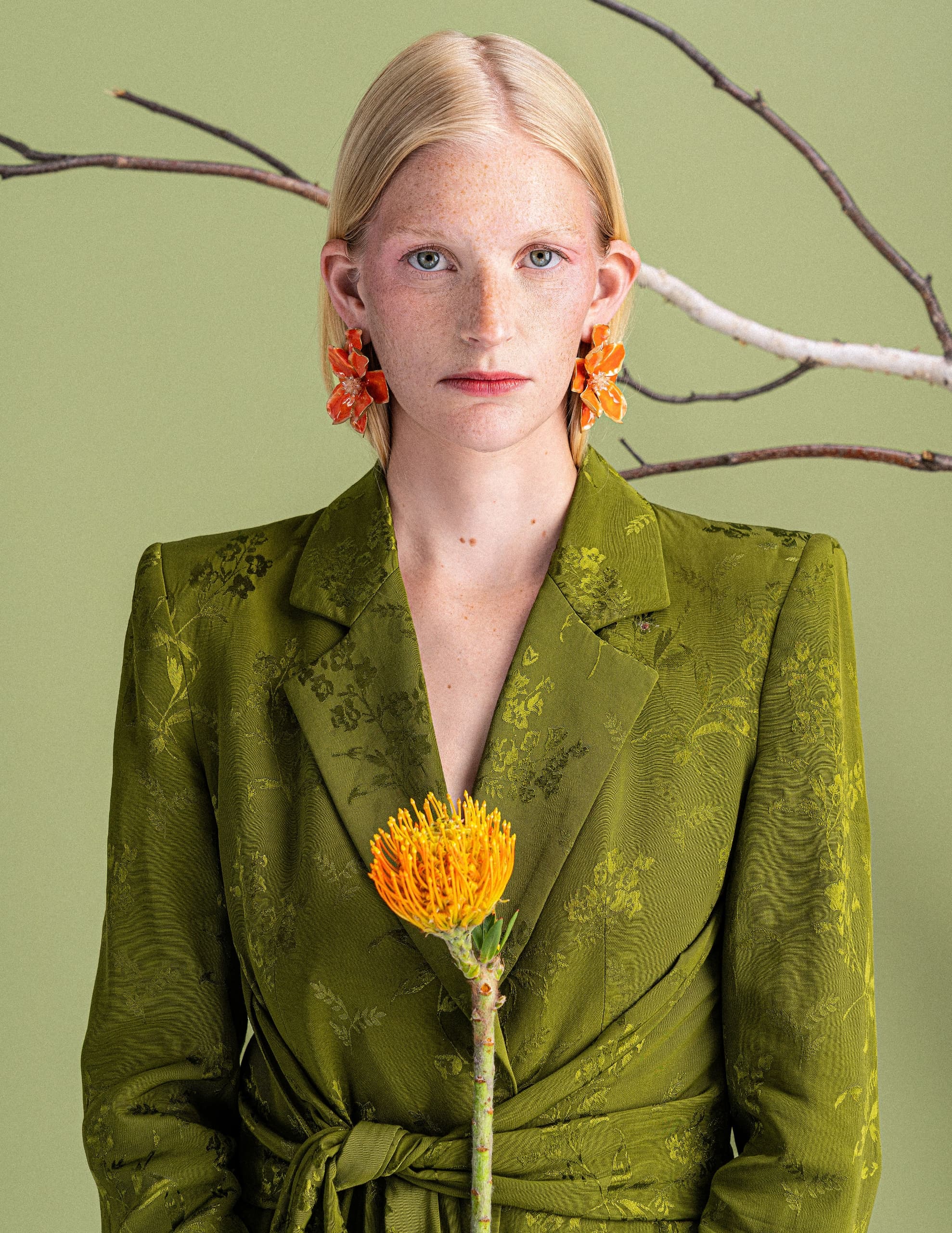 Woman in green suit with orange flower and earrings, set against a green backdrop.