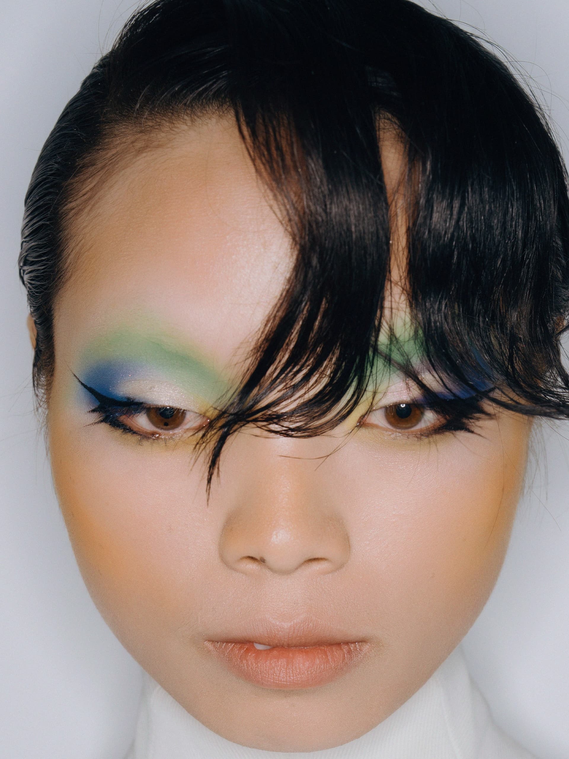Bold colorful eyeshadow with sleek hairstyle on woman wearing white high-neck top.