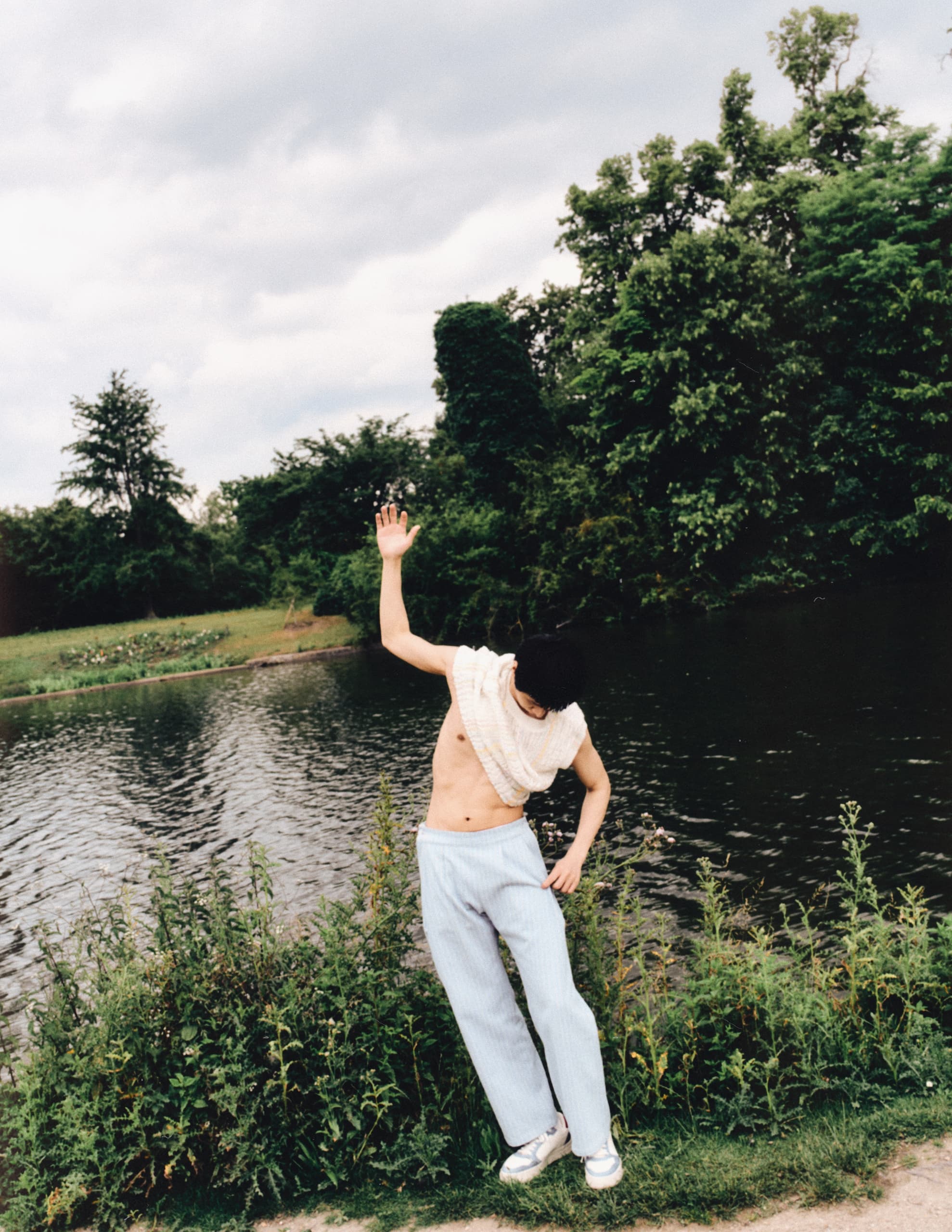 Person posing outdoors by a lake, surrounded by lush greenery and overcast sky.