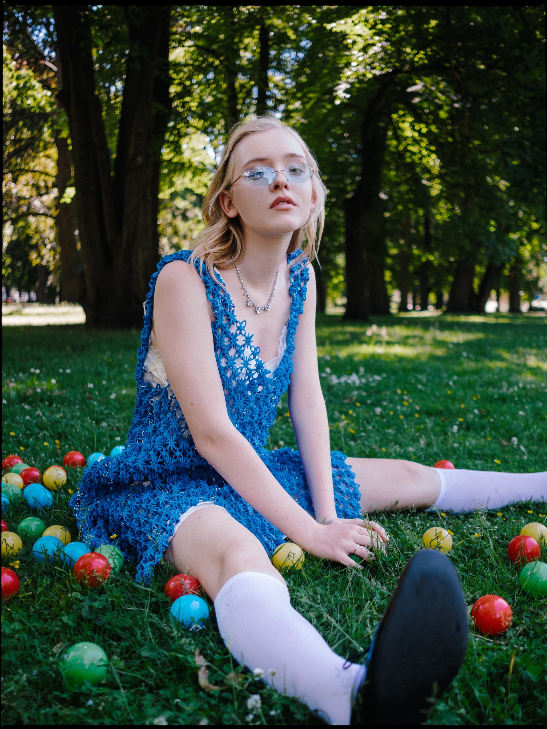 Person in blue dress sitting on grass, surrounded by colorful balls in a park.