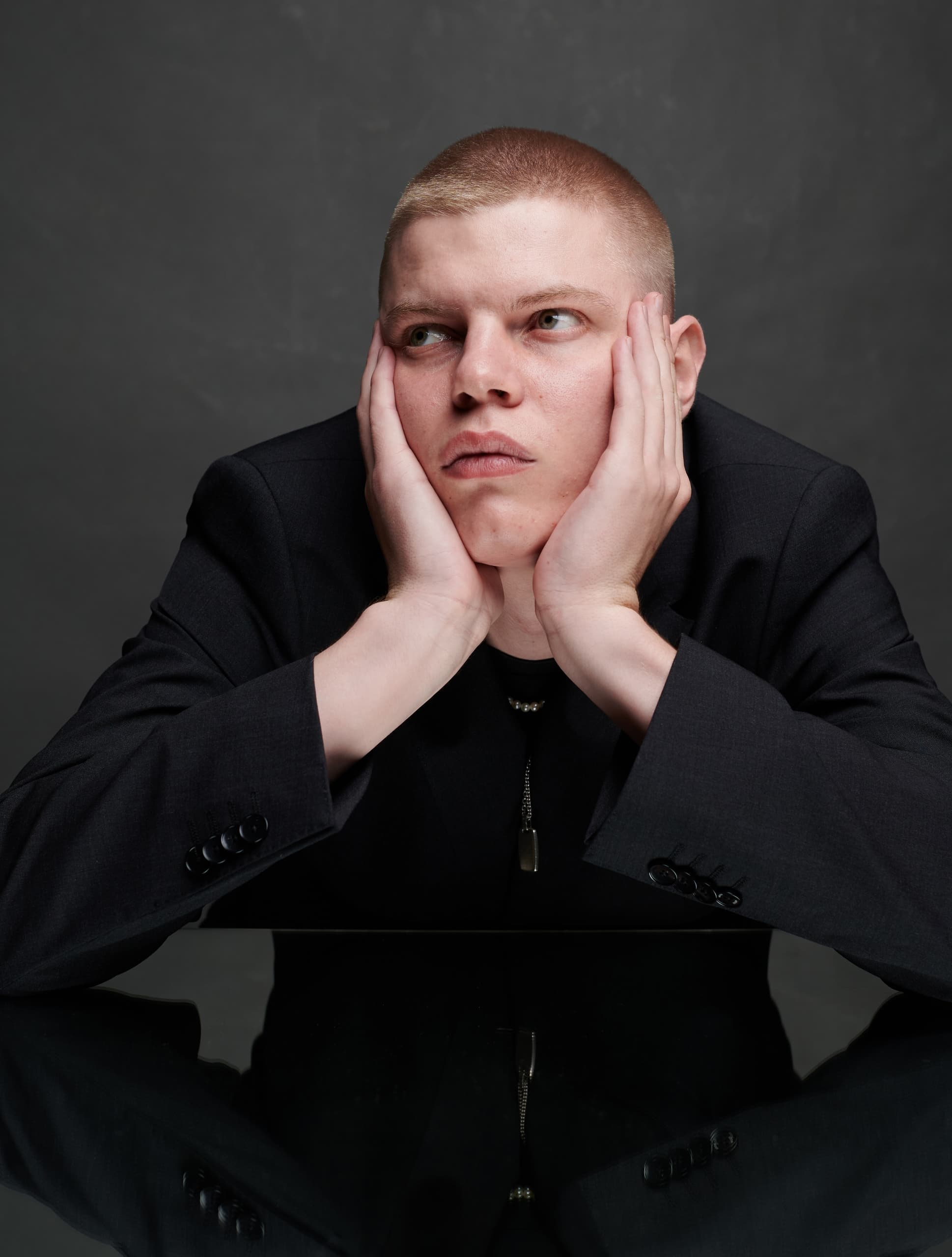 Man in a black suit looking contemplative with hands on cheeks against a dark background.