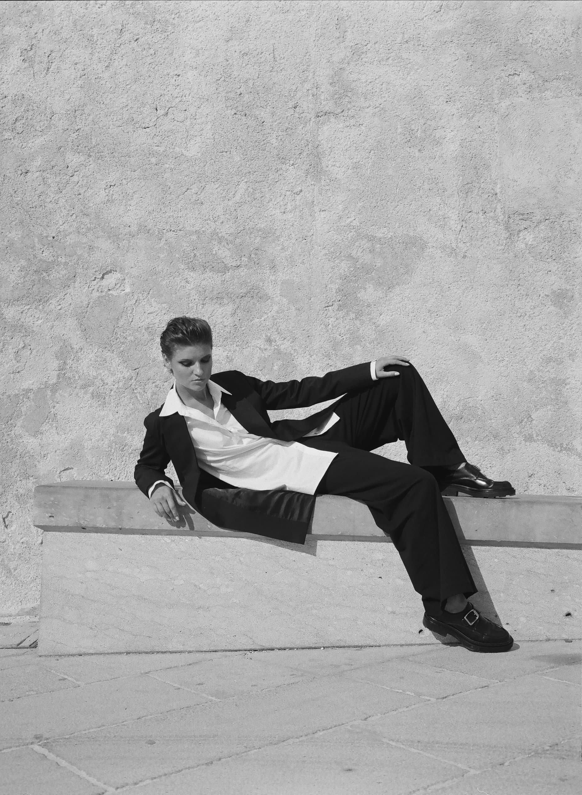 Person in black suit lounging on a bench against a textured wall.