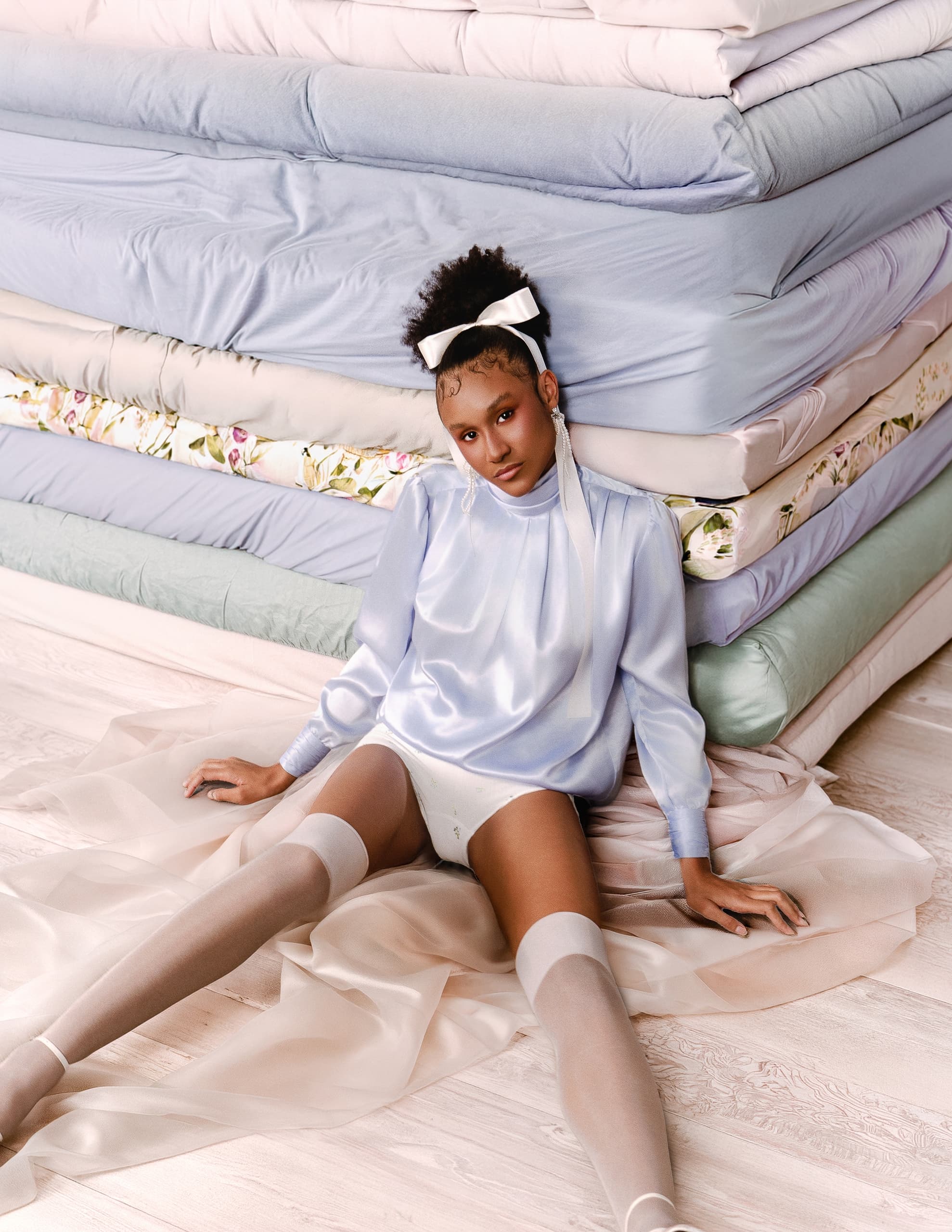 Young woman in elegant attire sits by tall stack of mattresses.