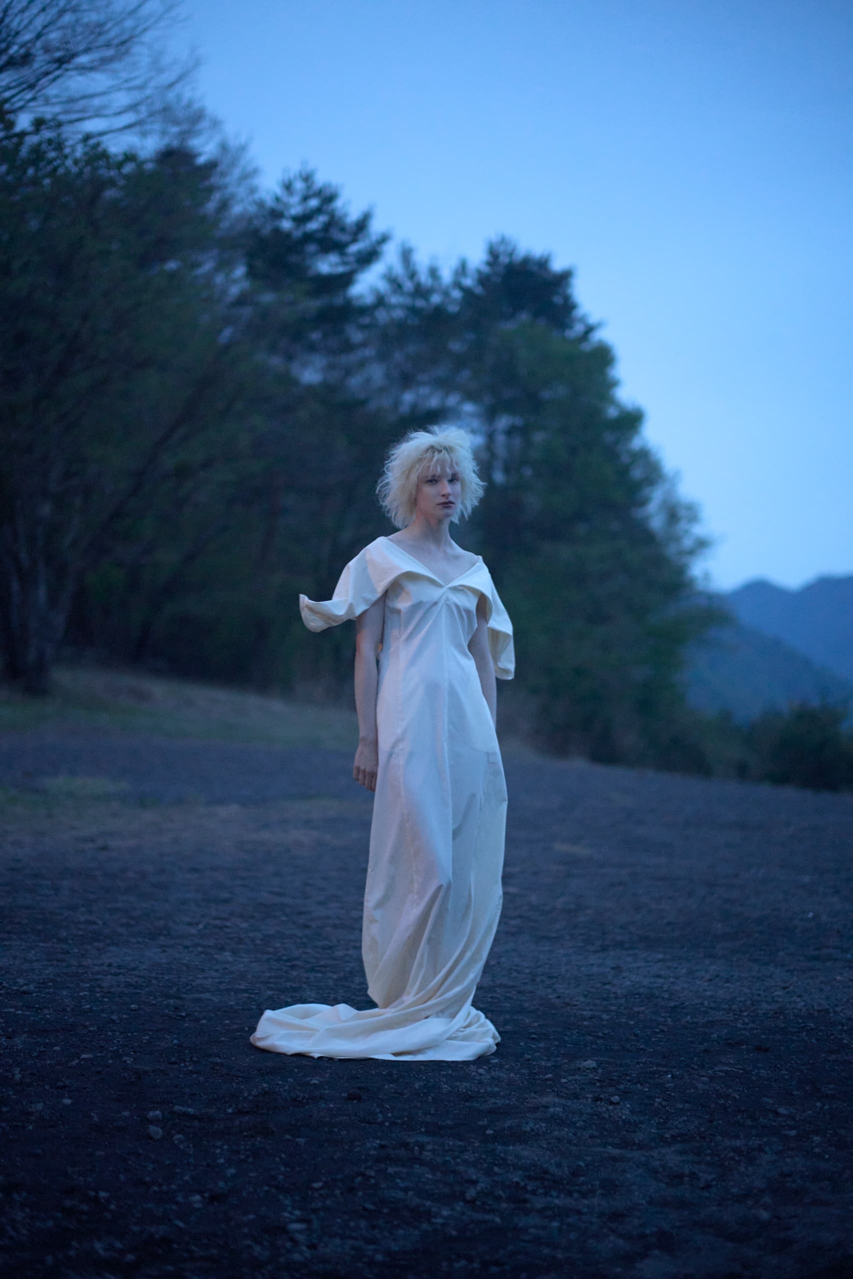 Person in a white gown standing outdoors at dusk in a serene natural setting.
