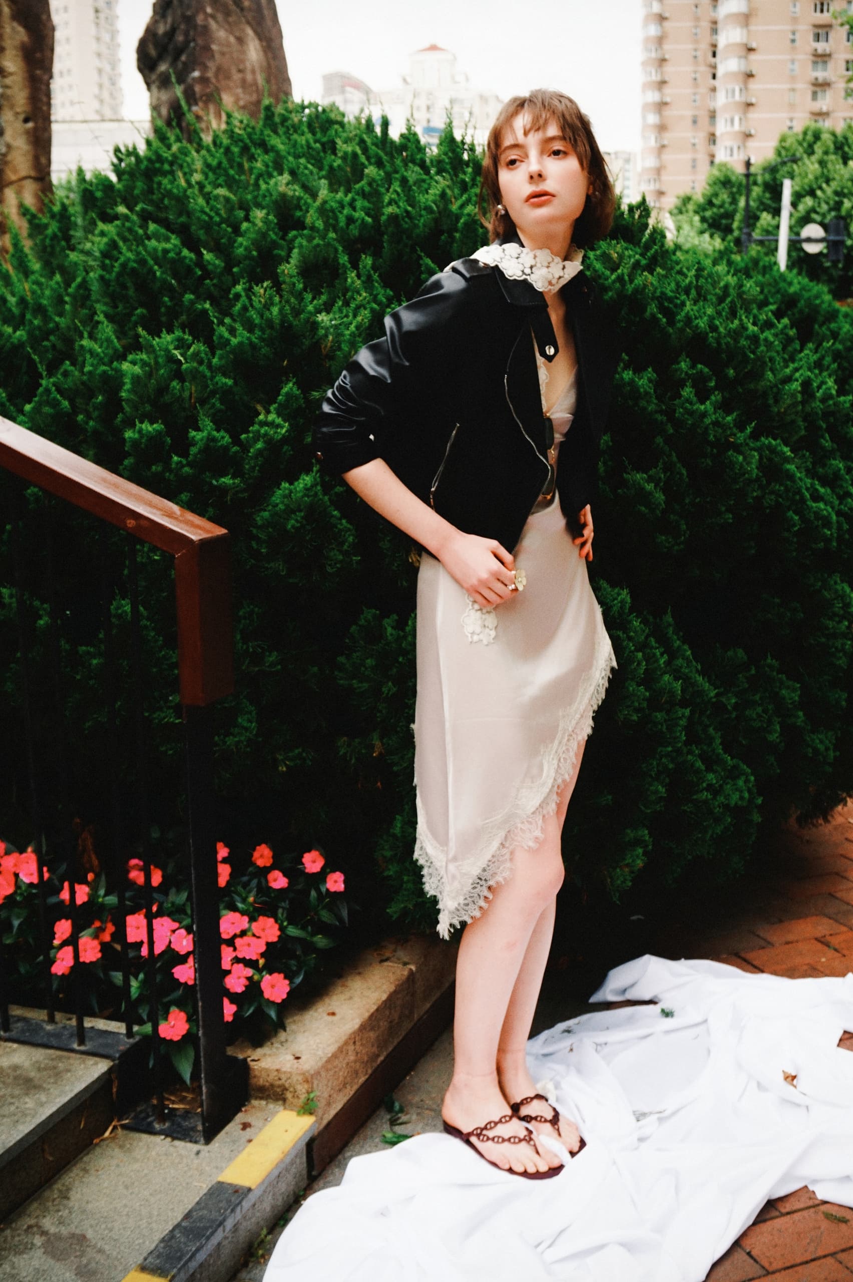 Fashionable woman in dress and jacket poses outdoors by greenery and flowers.