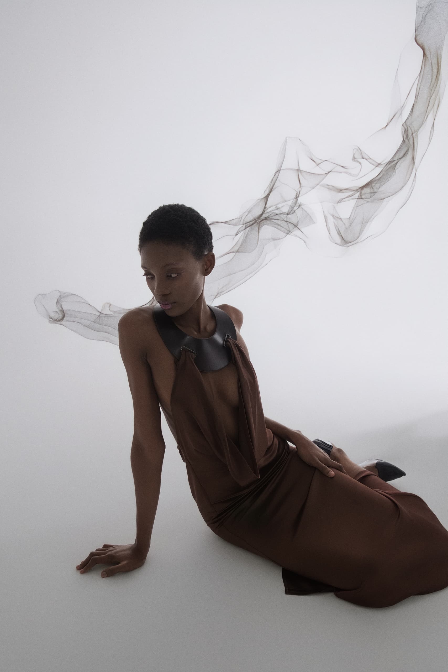 Elegant woman in a flowing brown dress with sheer fabric backdrop.