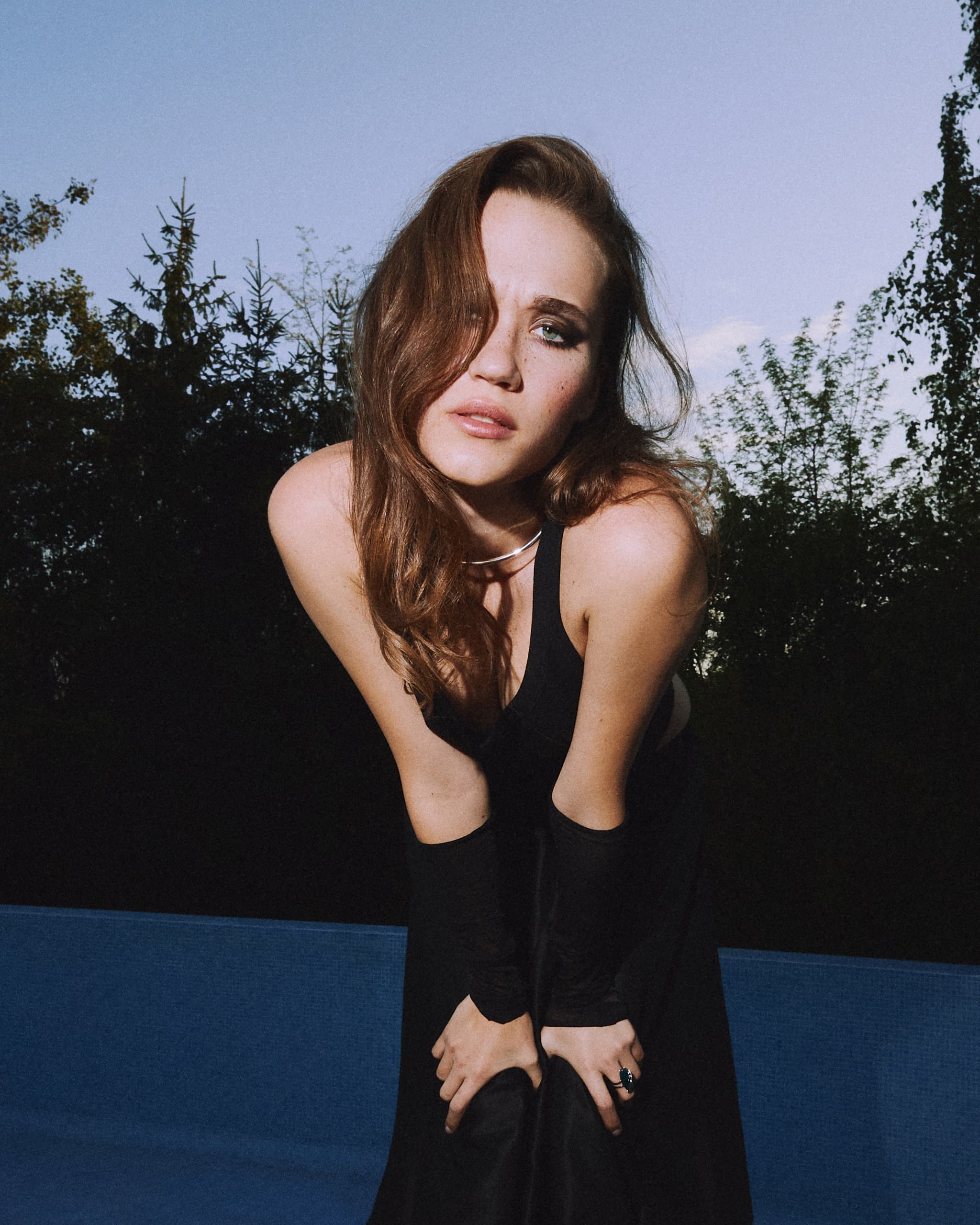 Woman in a black dress posing outdoors against a twilight sky and trees.