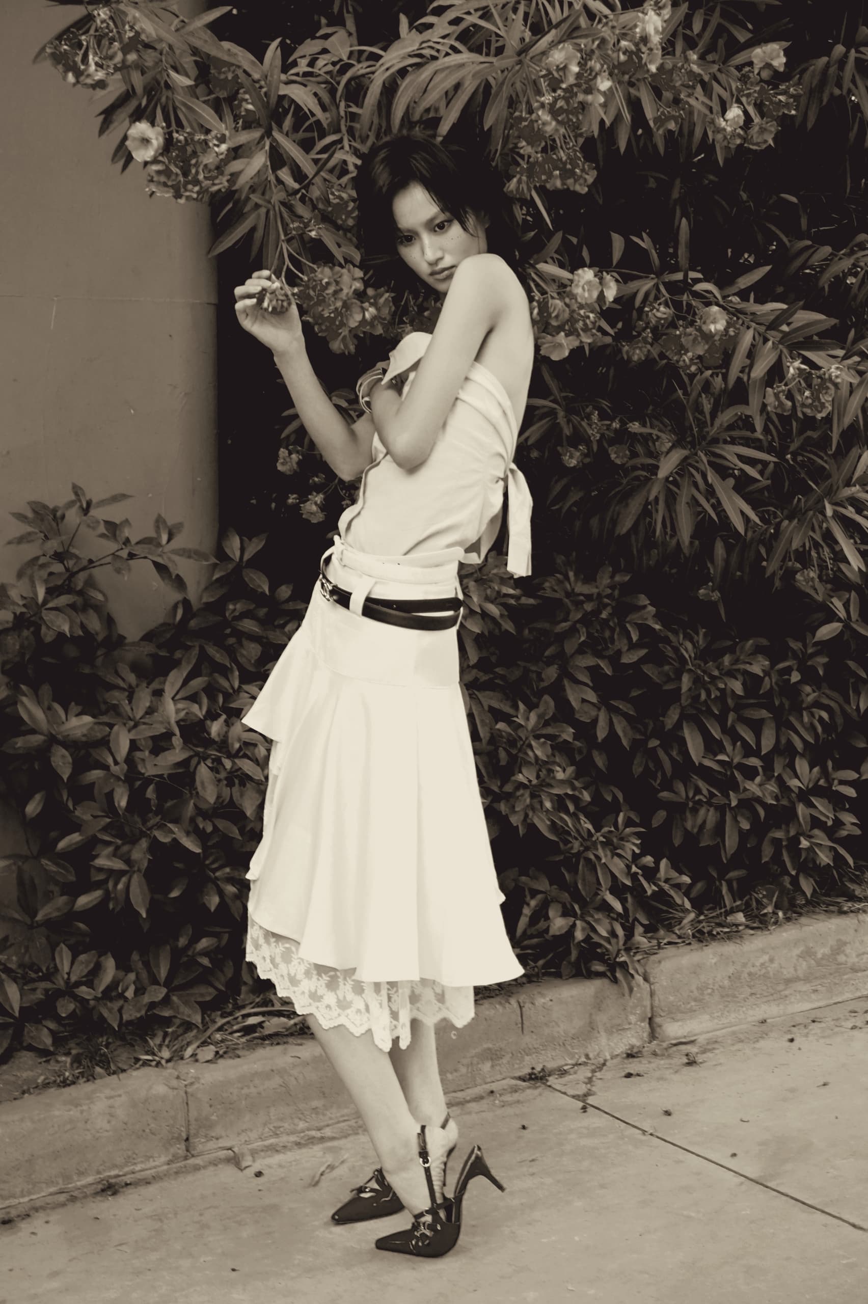 Woman in stylish outfit poses against leafy backdrop in artistic monochrome photo.