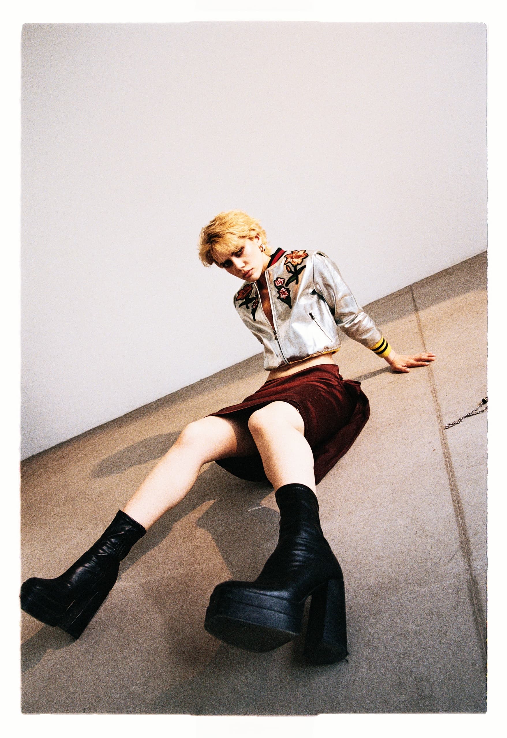 Fashionable model in embroidered jacket and platform boots seated on floor.