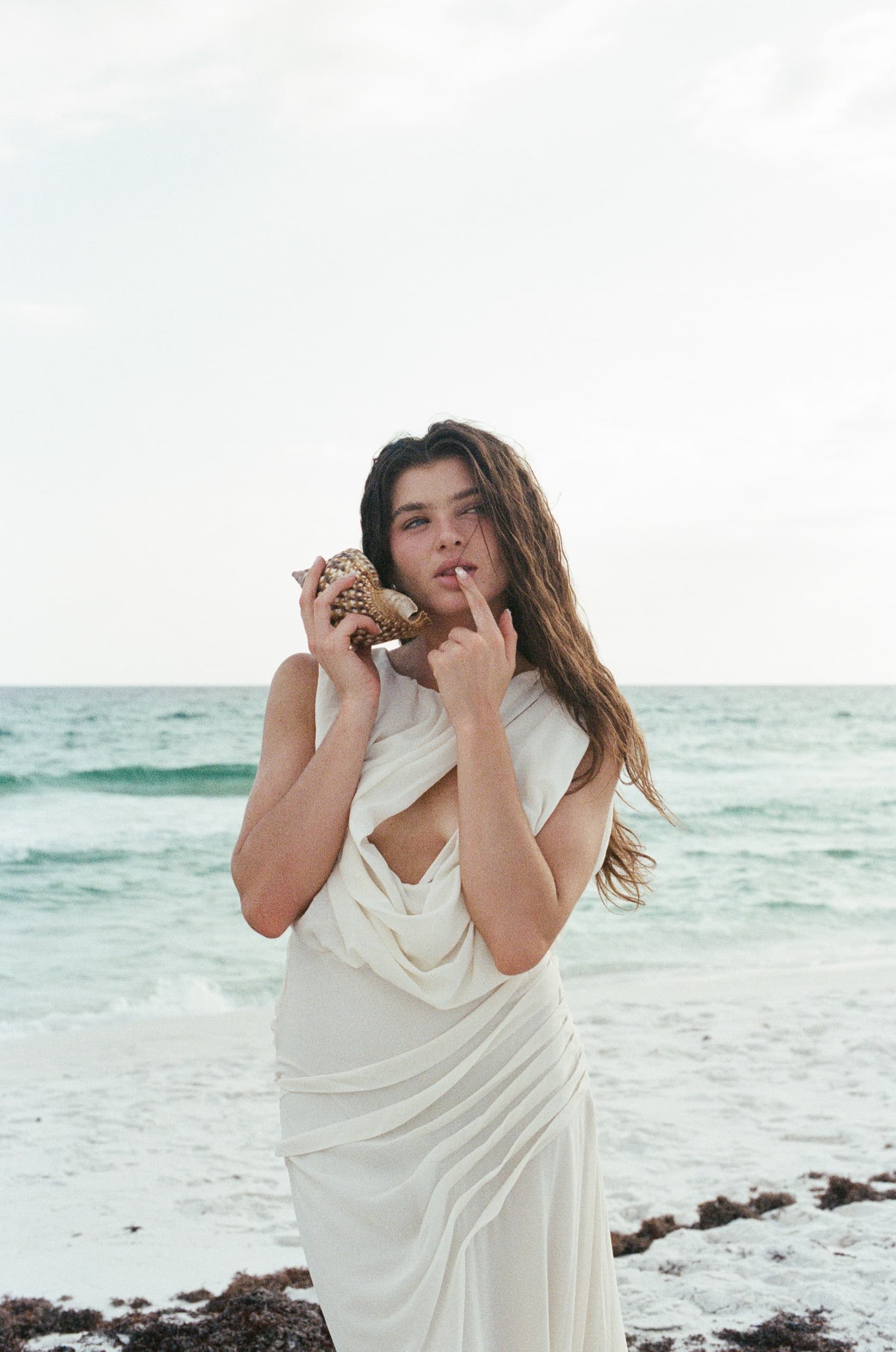 Woman in white dress holding a seashell by the ocean on a sandy beach.