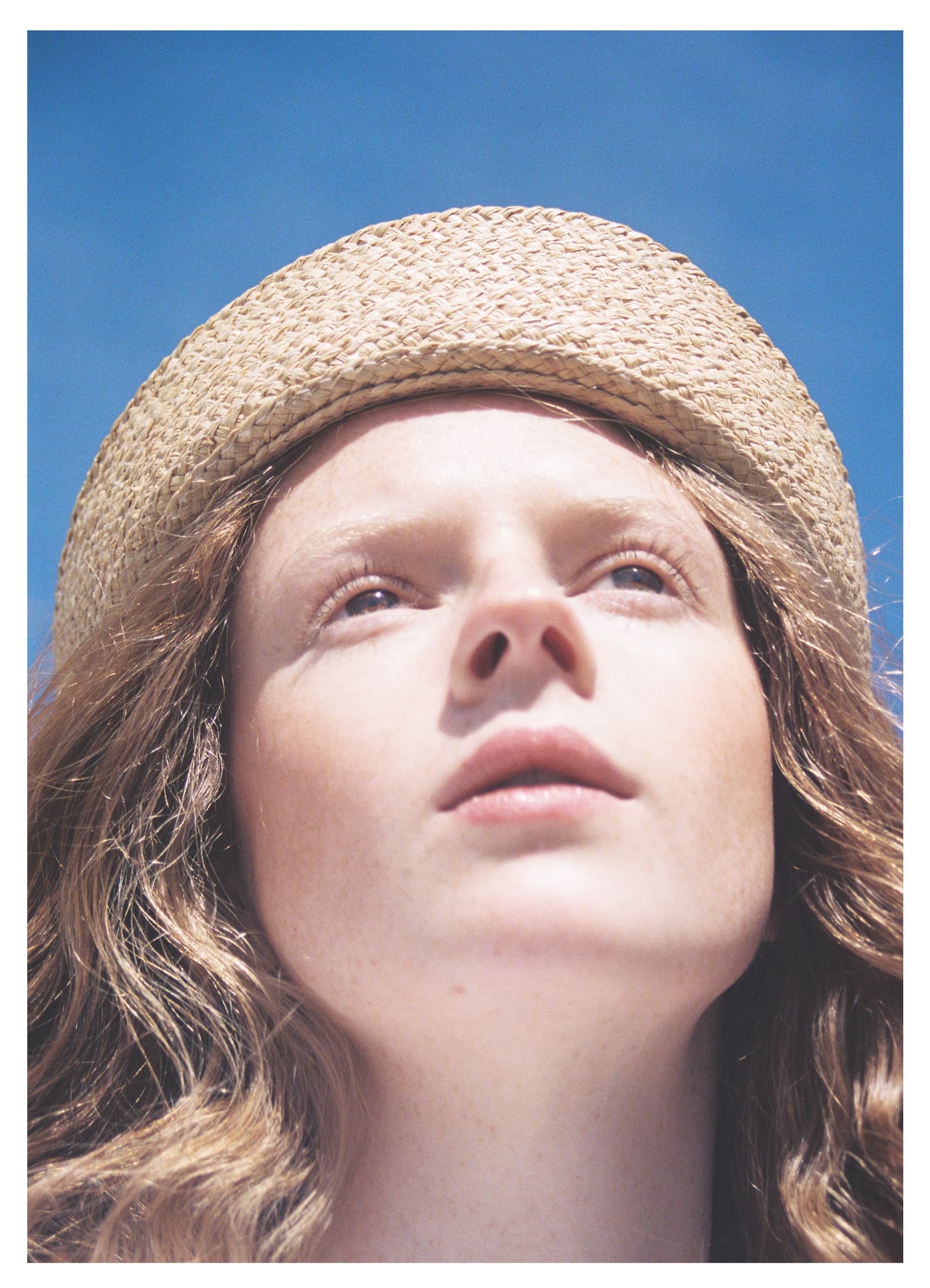 Woman with straw hat gazes up against blue sky background.