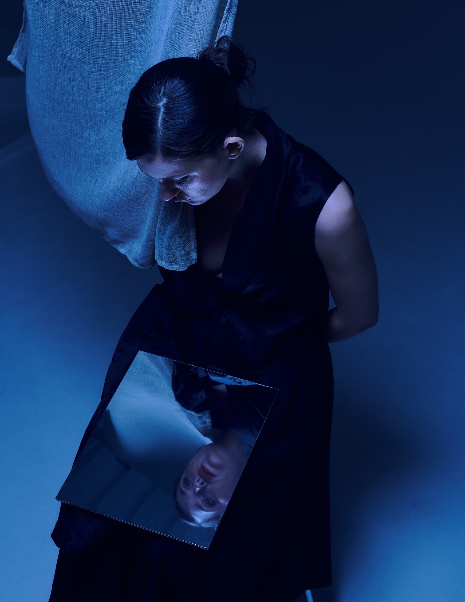 Woman in dark dress gazing down at mirror reflection in moody blue lighting.