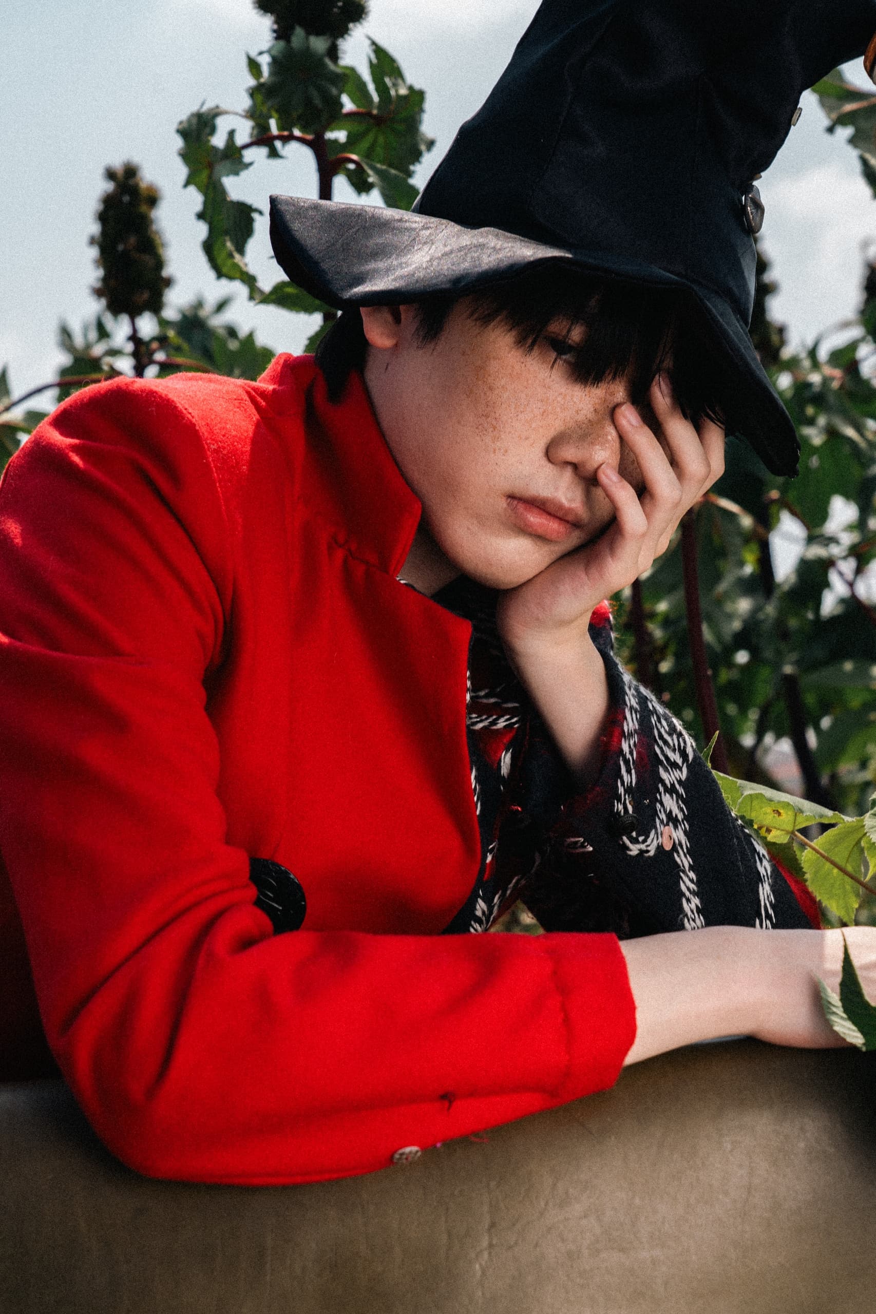 Person in a red coat and black hat resting with hand on face outdoors.